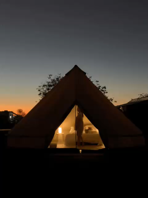 A large canvas expedition tent with an interior light on, set outdoors during sunset or dusk. The tent has an open entrance showing a bed inside, and a person standing in the doorway.