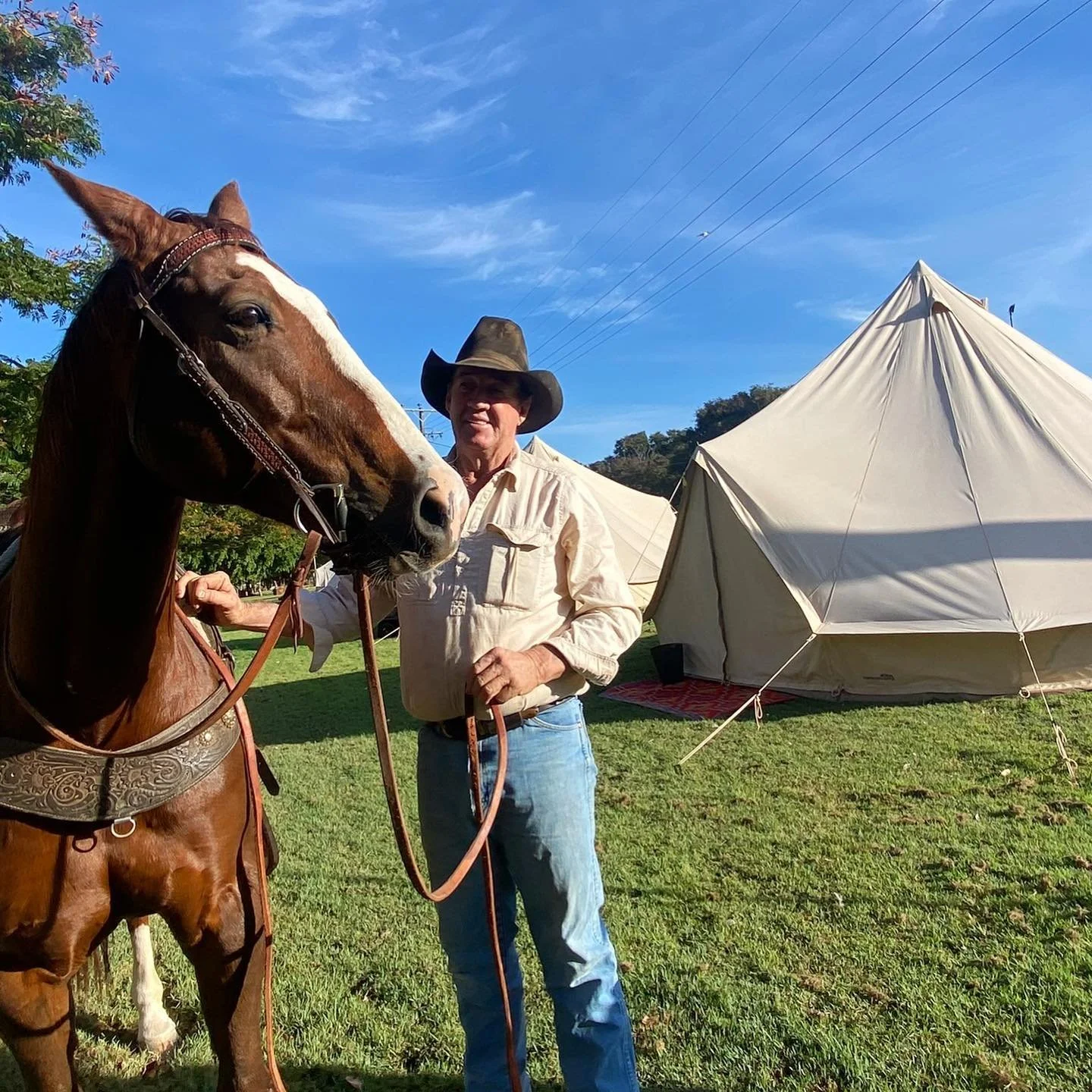A man wearing a cowboy hat and beige shirt standing with a brown horse, holding the horse's reins, outdoors on grass with a white camping tent and blue sky in the background.