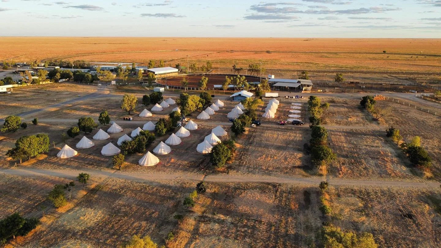 Aerial view of a rural farm camp with tents, trees, vehicles, and a few buildings in a farming area under a partly cloudy sky.