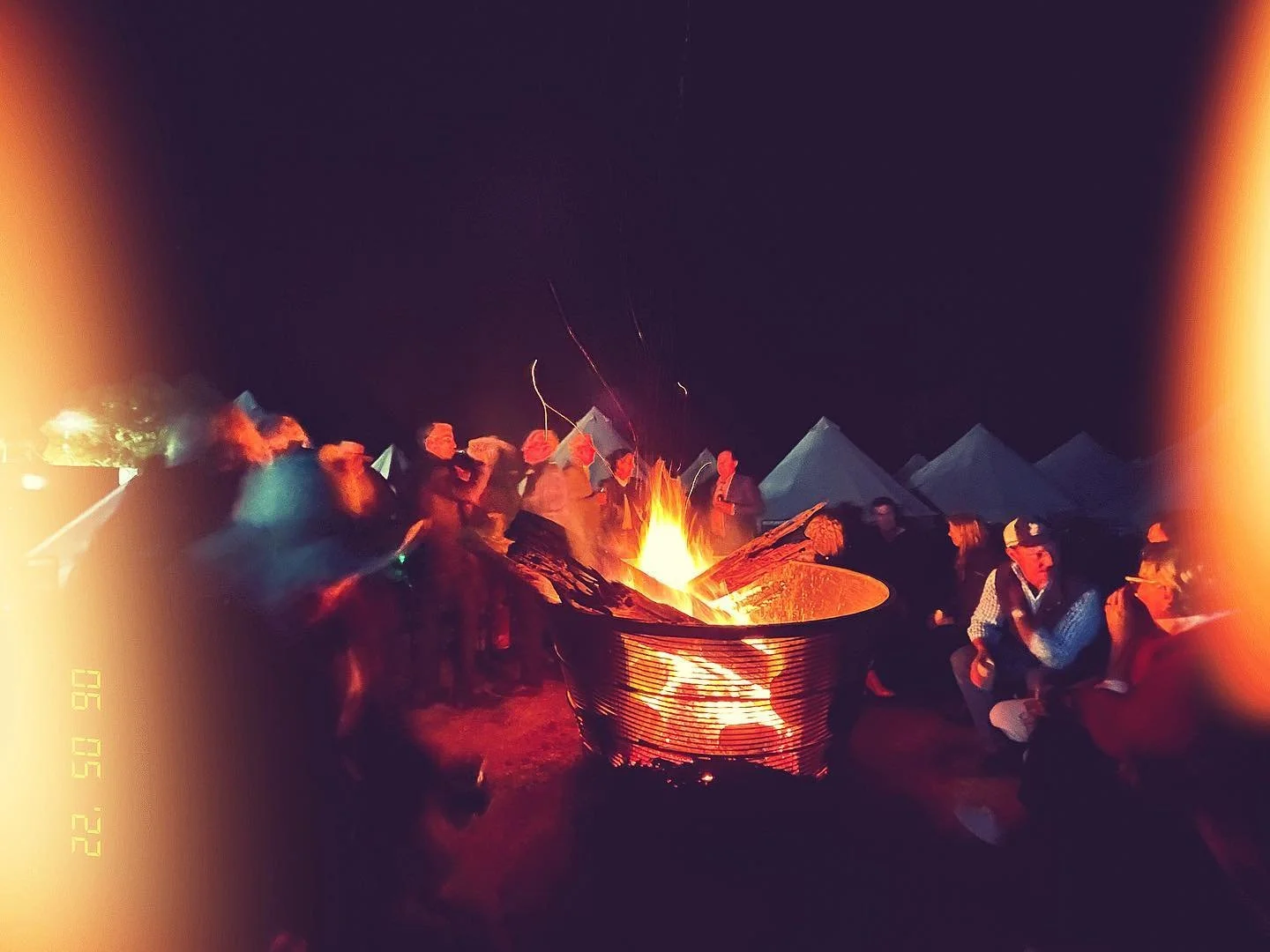 A group of people sitting around a campfire at night with tents in the background.