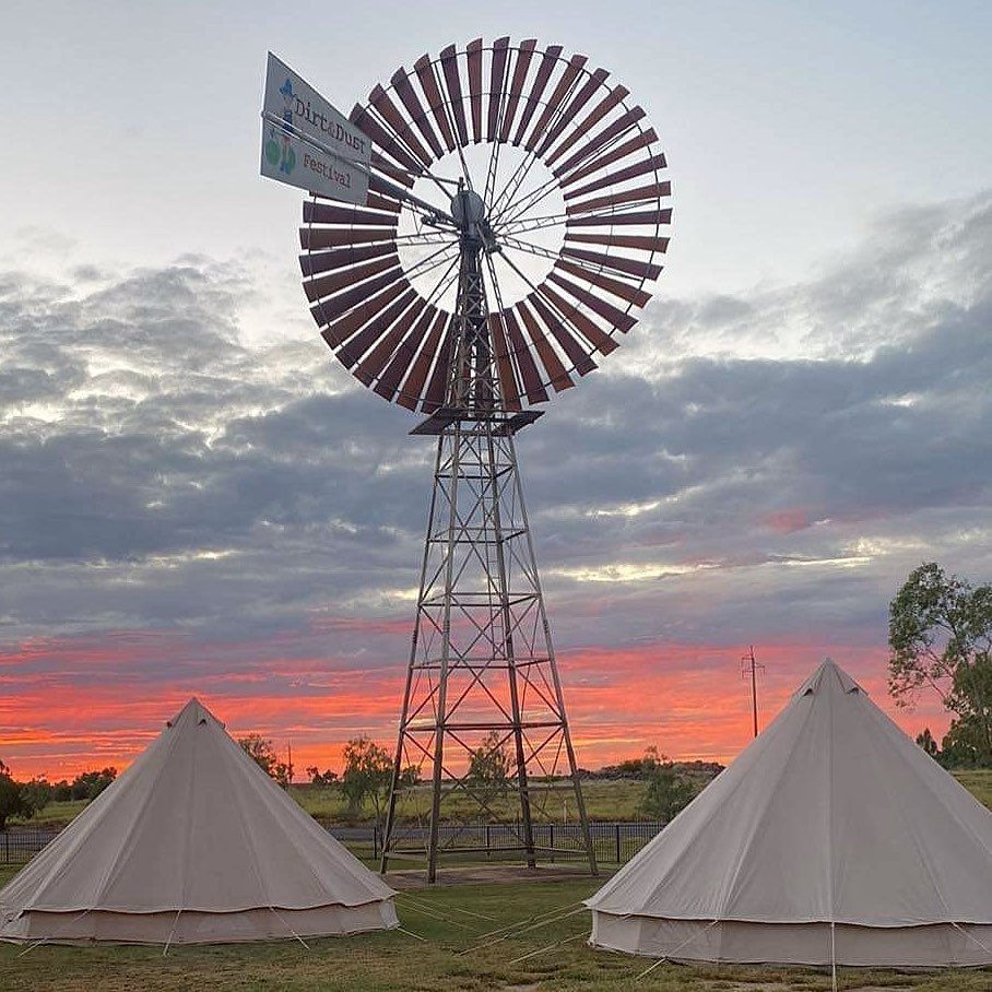 A windmill with a decorative circular blade design against a colorful sunset sky, with two white tents in the foreground.