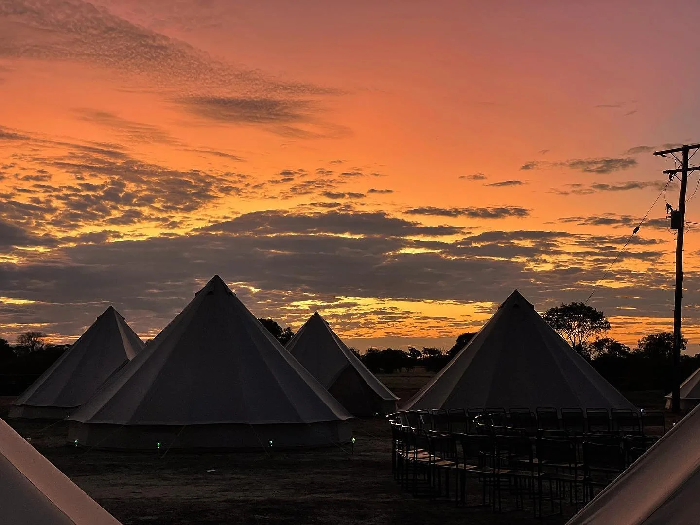 Sunset sky with orange and pink hues above dune-shaped tents set up outdoors with chairs in the foreground and electrical pole to the right.