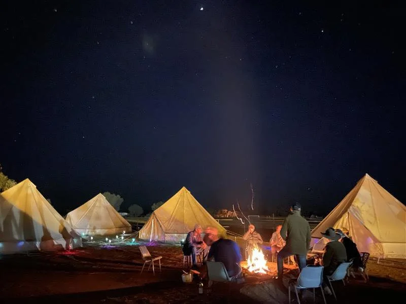 People sitting around a campfire at night, with tents set up on a field under a starry sky.