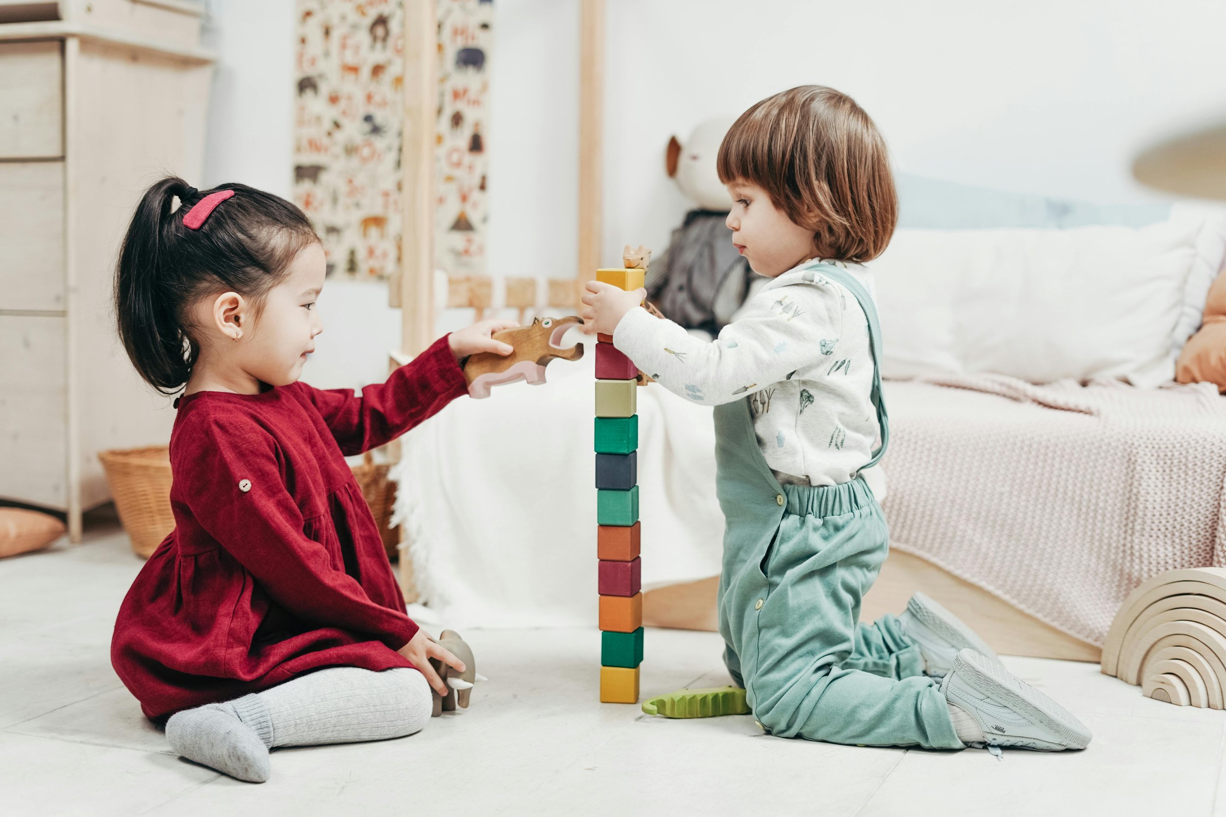 Two young children, a girl in a red dress and a boy in teal overalls, are playing together indoors. The girl is sitting on the floor and holding a wooden dinosaur toy, while the boy, kneeling, is stacking colorful blocks on a tall tower. Behind them is a bed with a beige blanket and pillows, and a decorative wall hanging with animal illustrations.
