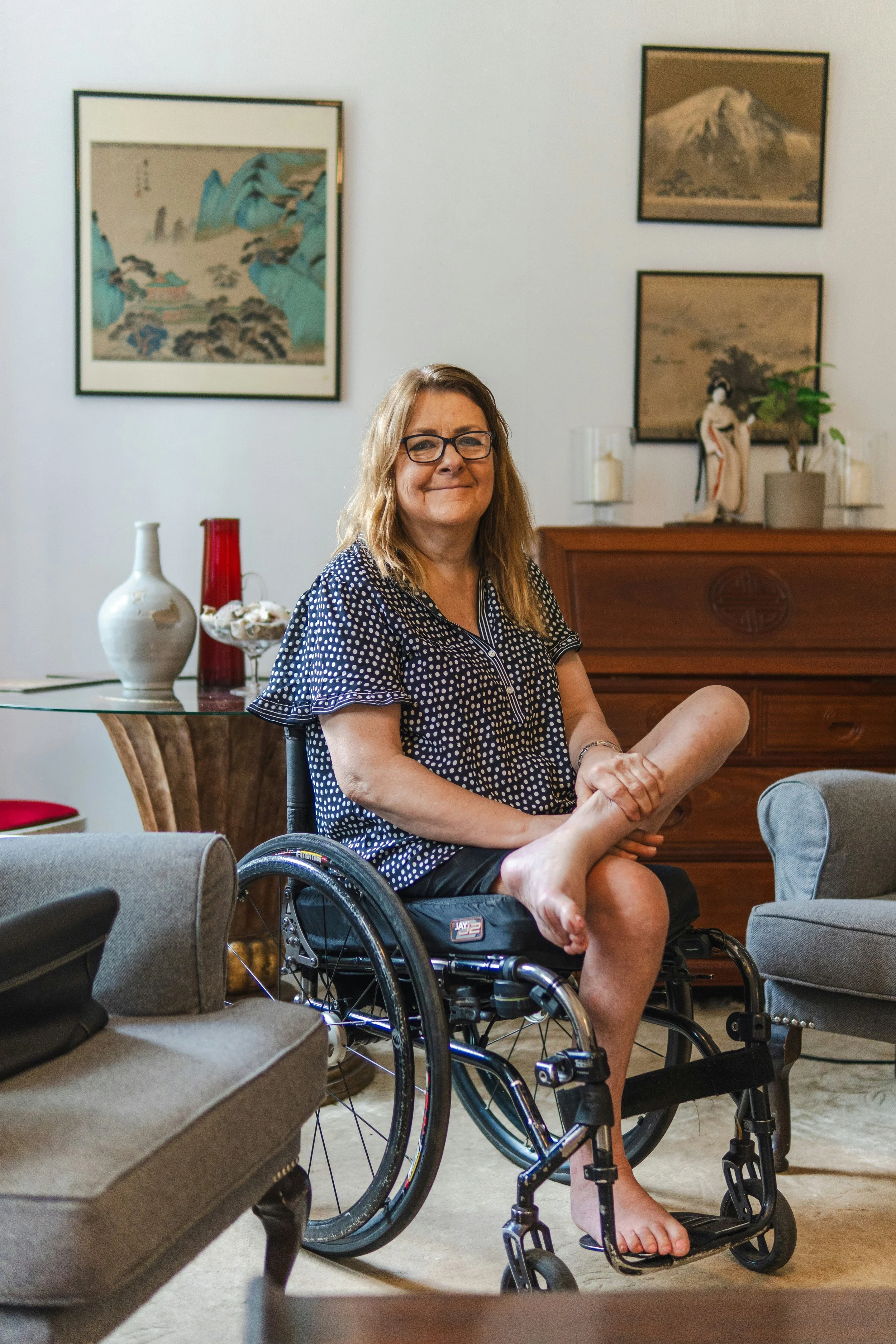 A woman in a wheelchair sitting in a living room with framed artwork on the wall, a wooden sideboard with decorative items, and a glass table with vases.
