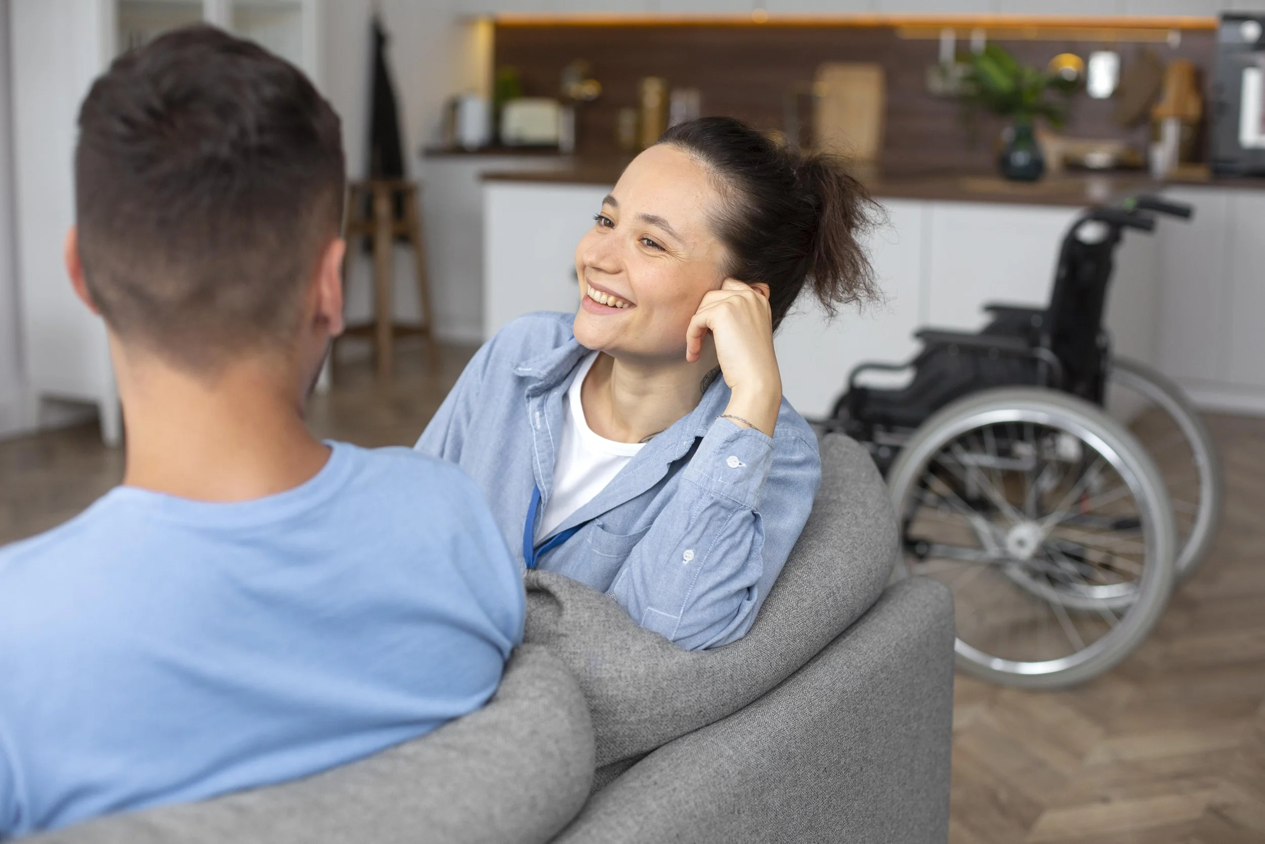 A woman smiling and looking at a man with his back to the camera, sitting on a gray couch in a modern kitchen. A wheelchair is visible in the background.