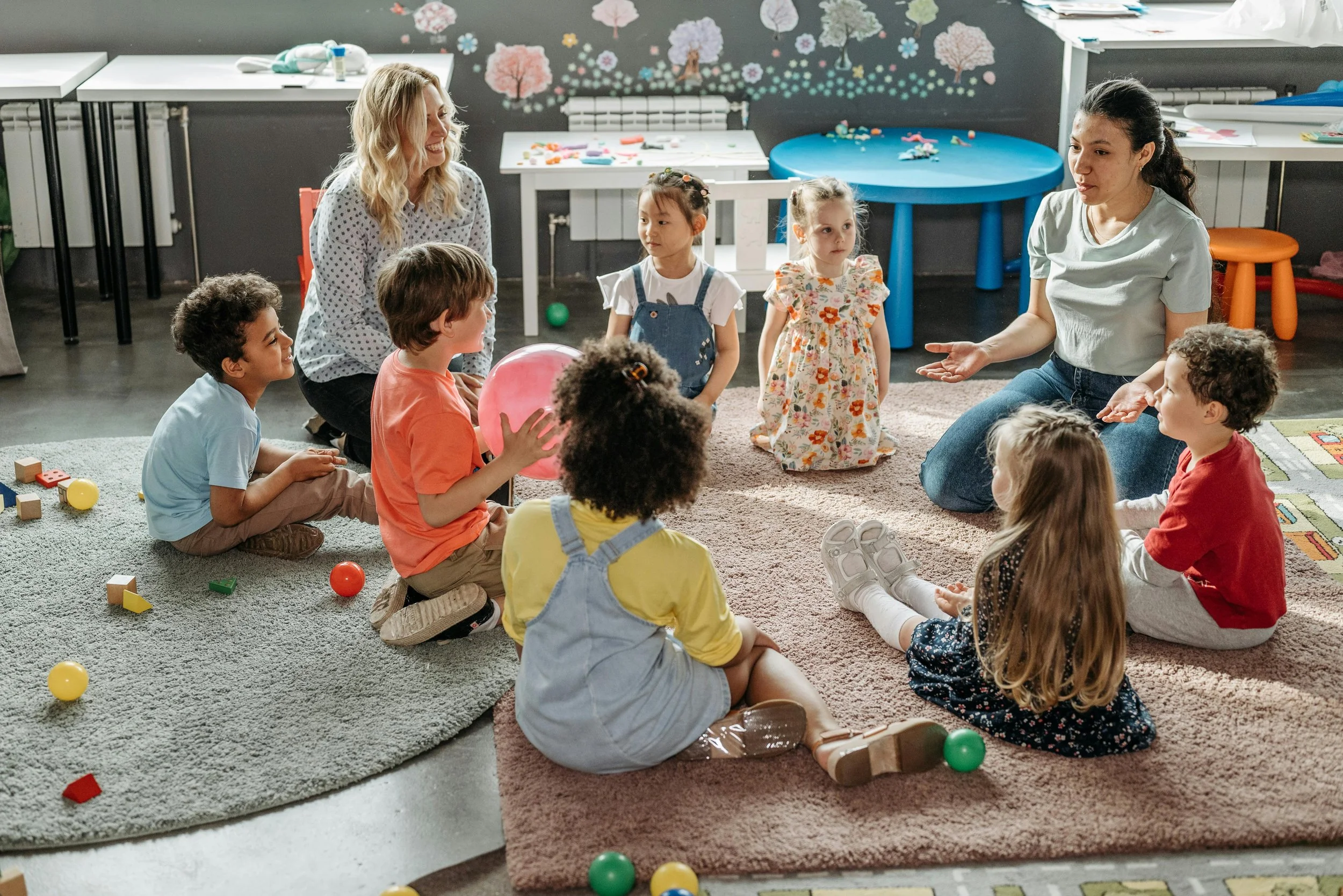 A group of young children sitting on a carpeted floor in a classroom, engaged in a discussion with two adult women. There are colorful toys and educational materials around them, and the classroom walls are decorated with pink and white cherry blossom trees.