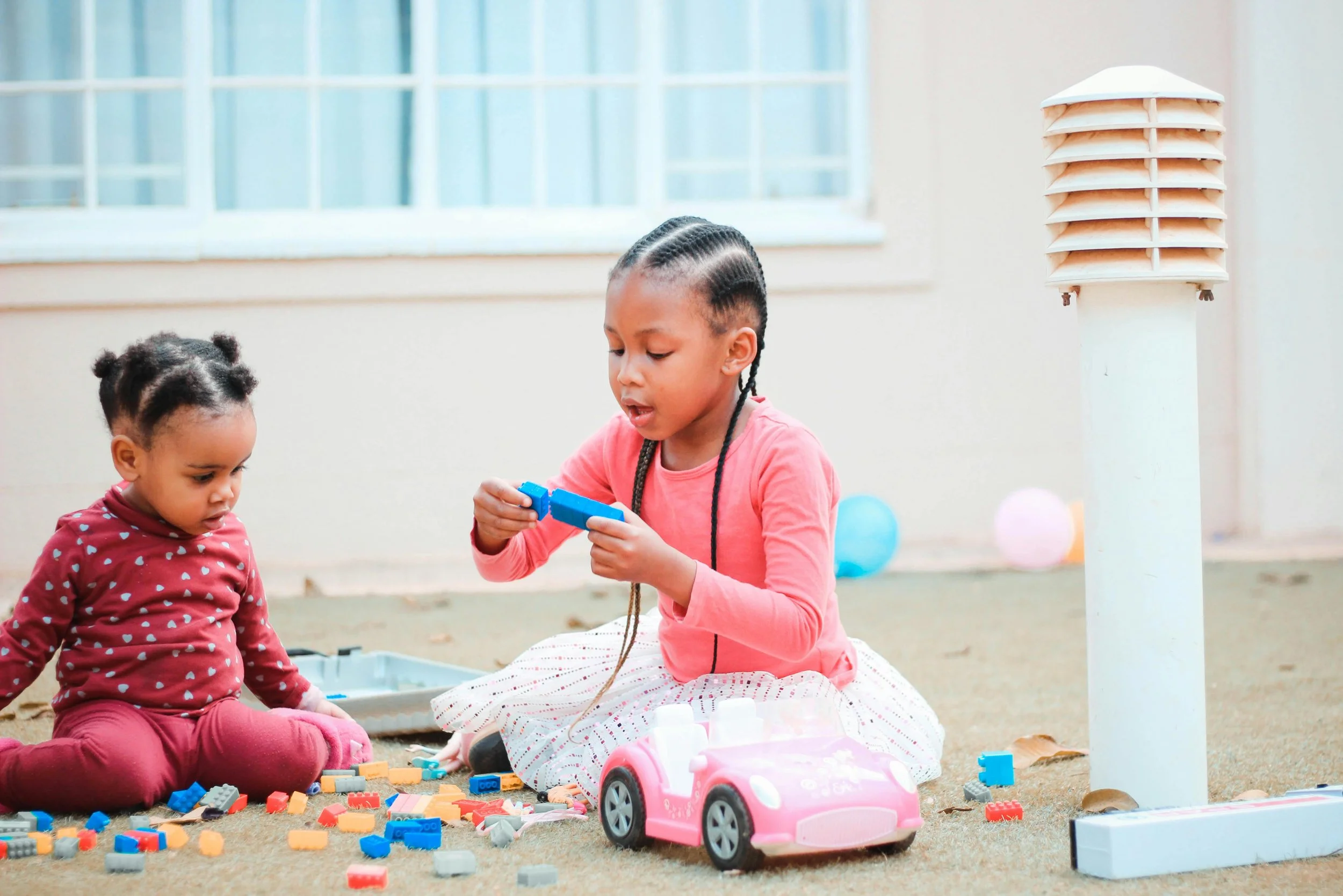Two young girls playing with building blocks and a toy pink car outdoors near a beige wall with window. One girl is holding a blue block, and the other girl kids are sitting on the ground surrounded by colorful blocks.