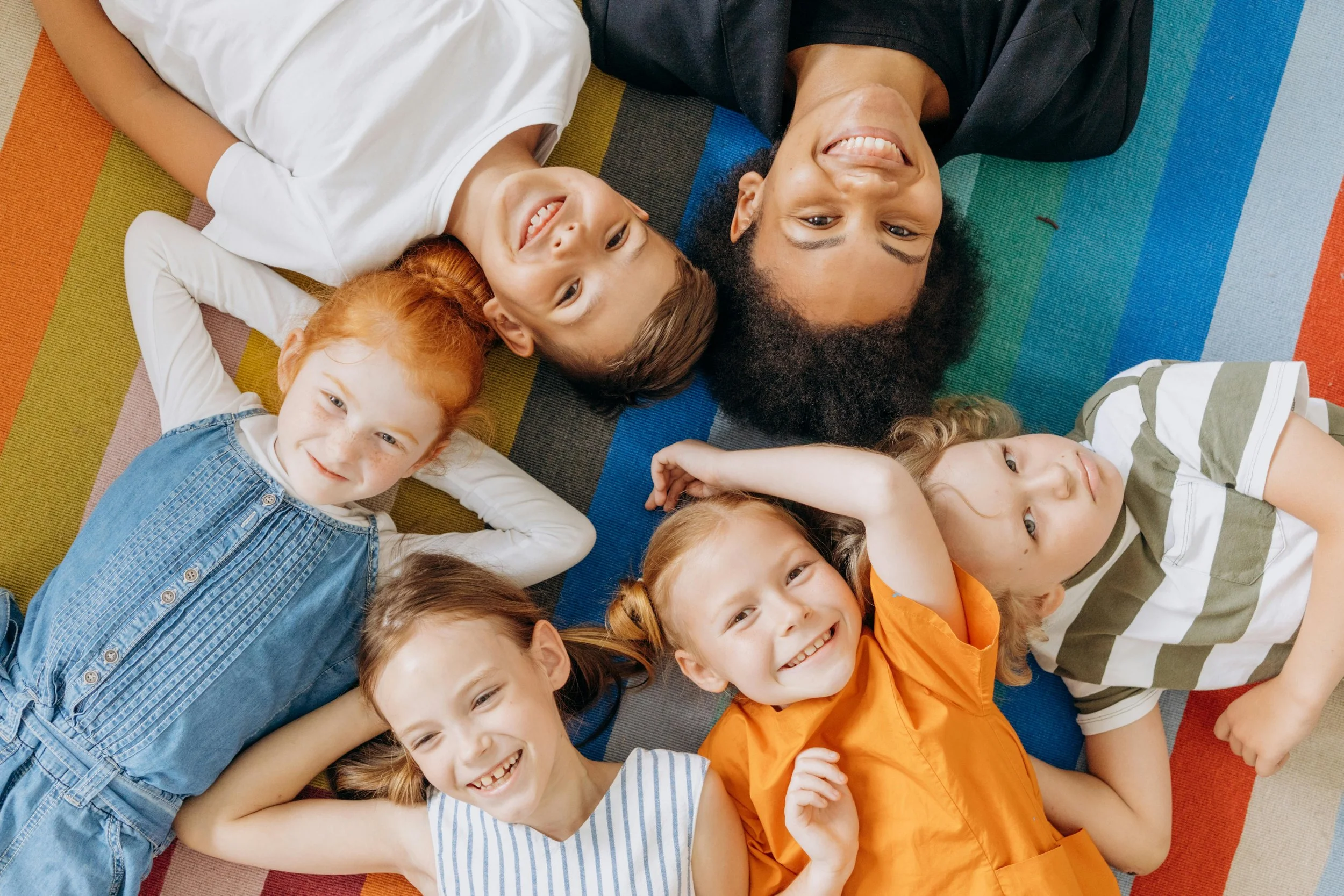 A diverse group of children and an adult lying on a colorful striped rug, smiling at the camera.