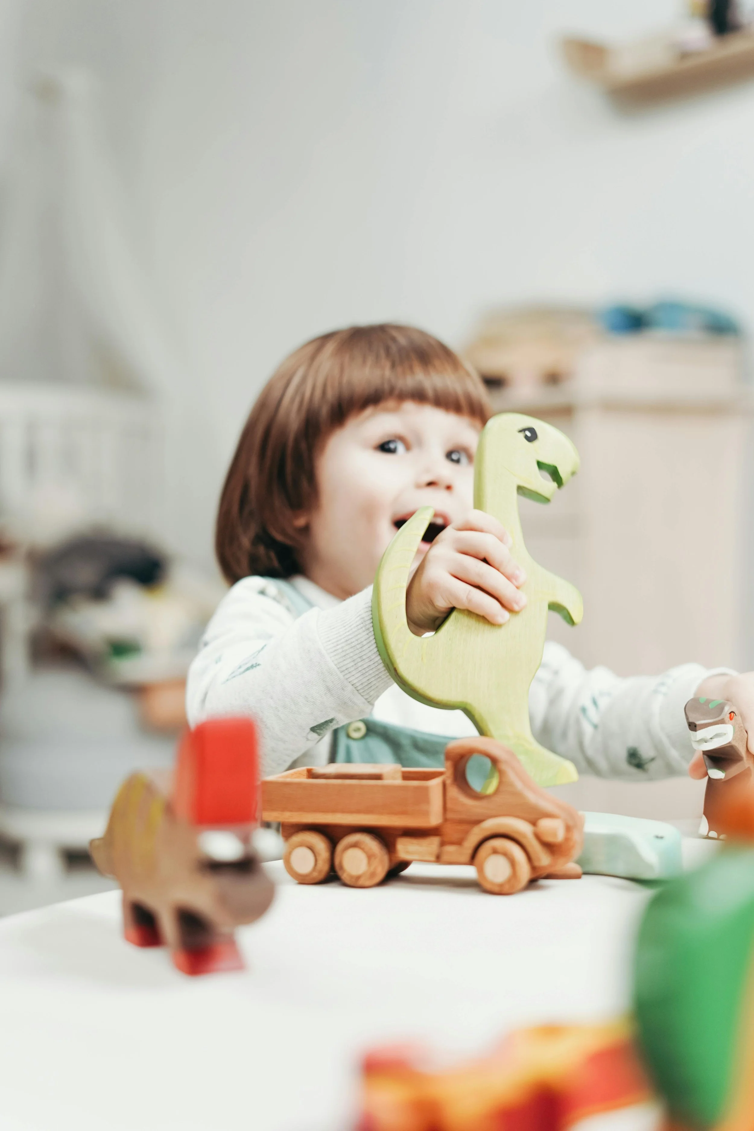 Child playing with wooden toys, including a dinosaur and a toy truck, at a table in a room.