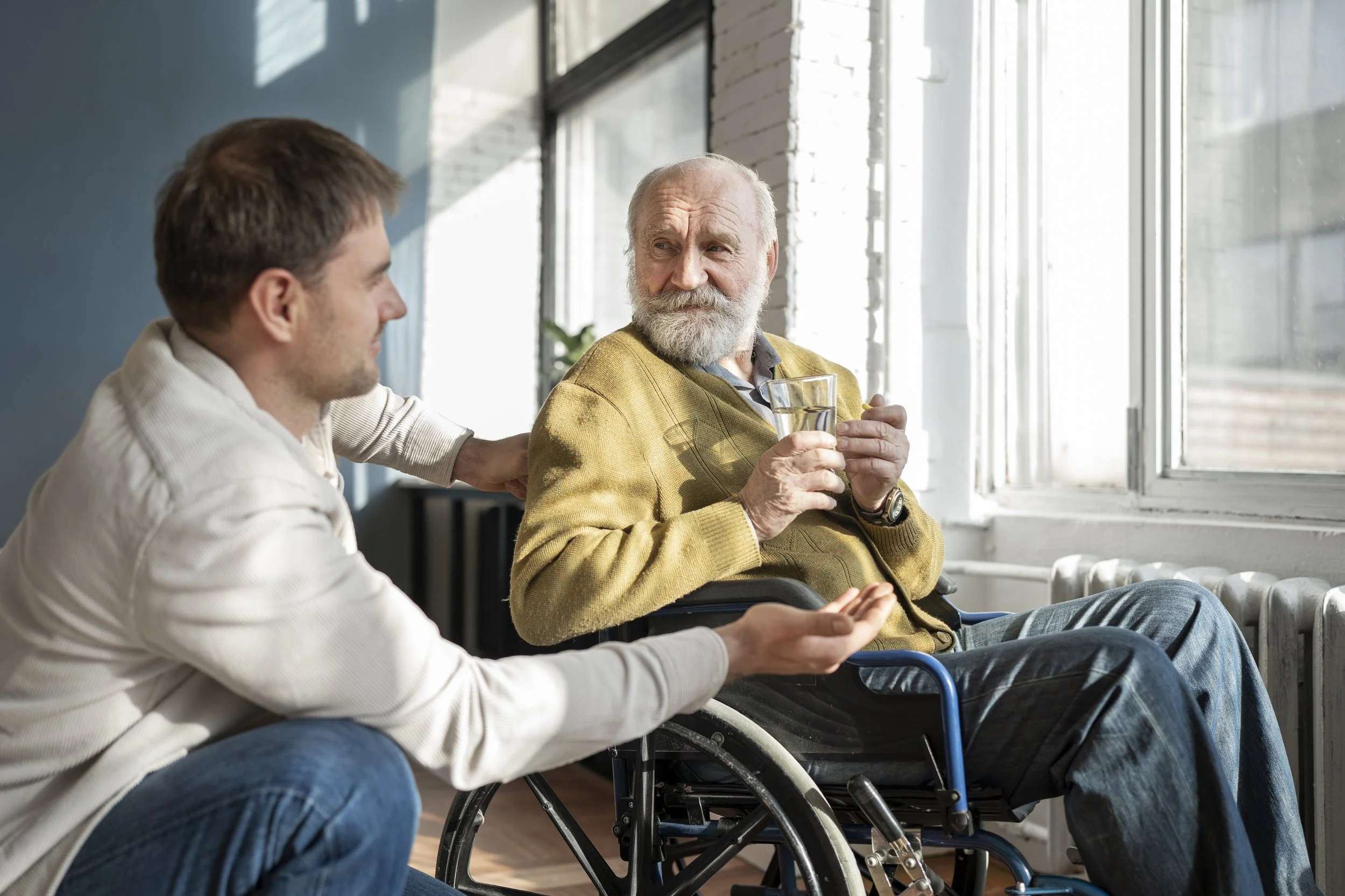 An elderly man with a white beard in a yellow sweater and gray pants is holding a glass of water and sitting in a wheelchair, engaging in conversation with a young man with brown hair in a white shirt, in a room with large windows and white brick walls.