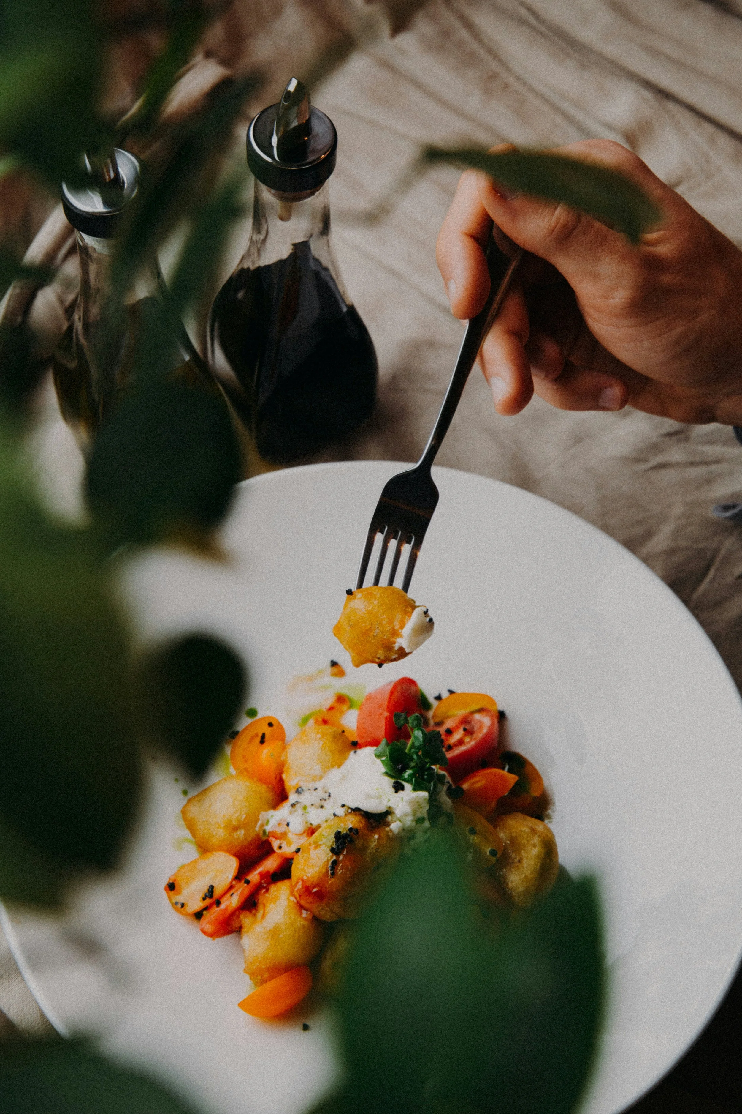 Une assiette contenant un plat gastronomique avec des légumes colorés, une sauce blanche, et une garniture noire. Un bras tenant une fourchette pique un morceau de l'assiette. Autour, des bouteilles de sauce ou d'huile. L'image est prise en gros plan, avec une mise en scène élégante.