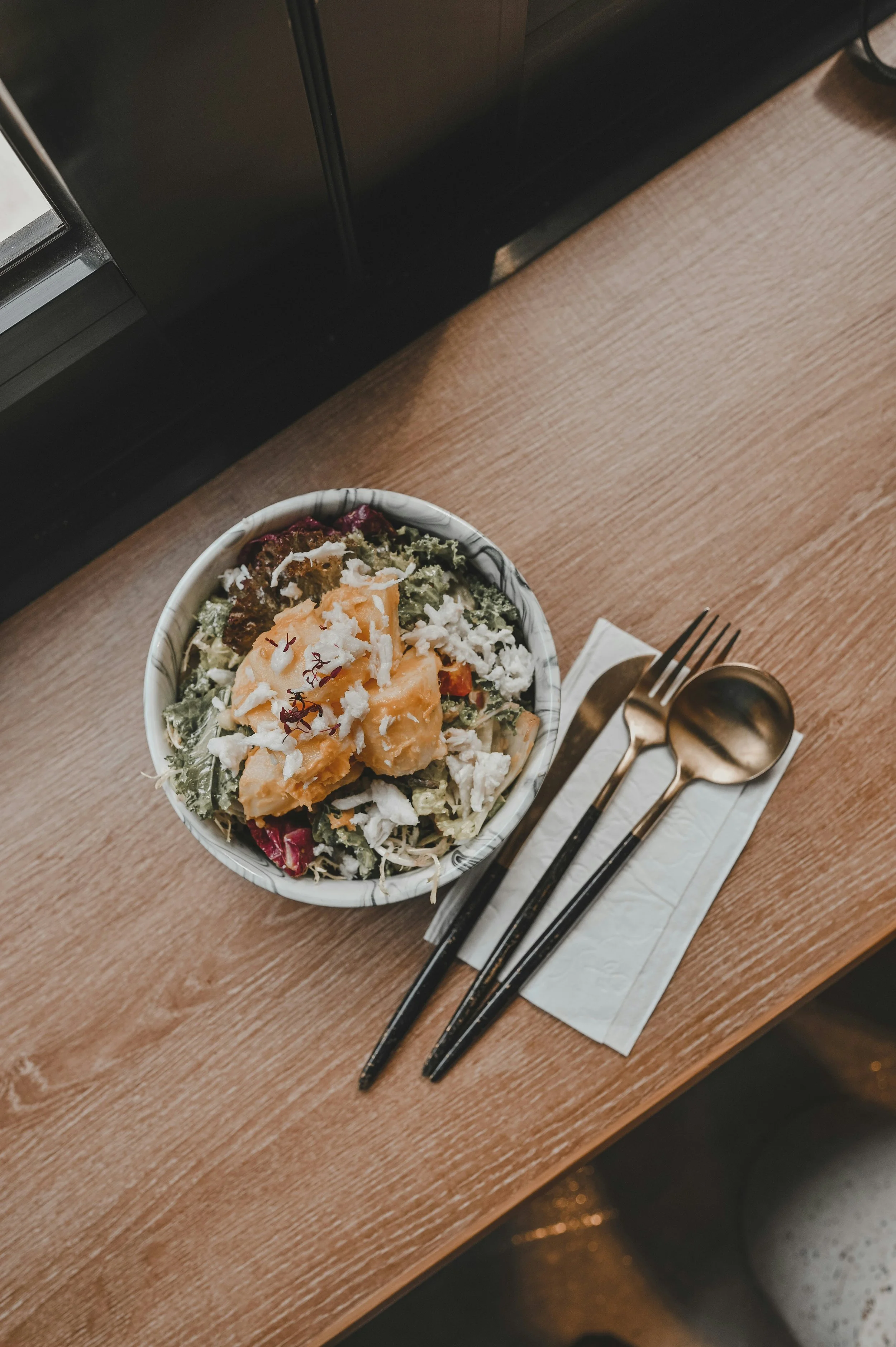 Salade dans un bol en céramique avec un décor de marbre, garnie de légumes, fromage et morceaux de melon, placé sur une table en bois avec une fourchette, une cuillère et une petite cuillère dorée posées sur un serviette en papier blanc.