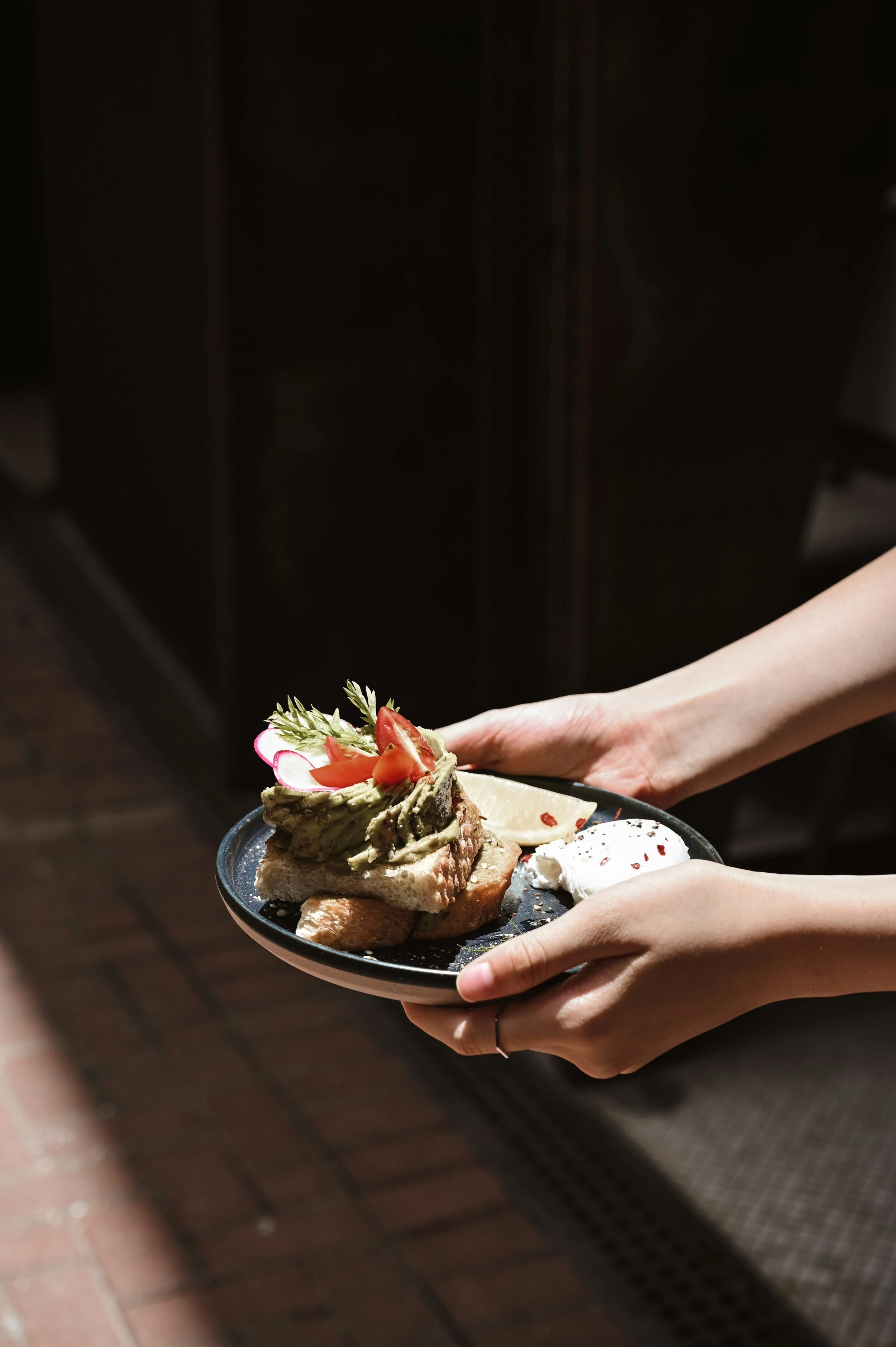 Une personne tient une assiette avec un plat de nourriture, comprenant du pain, une tartine garnie d'avocat, de tomates, des radis, une tranche de fromage et un œuf poché, dans un environnement intérieur.