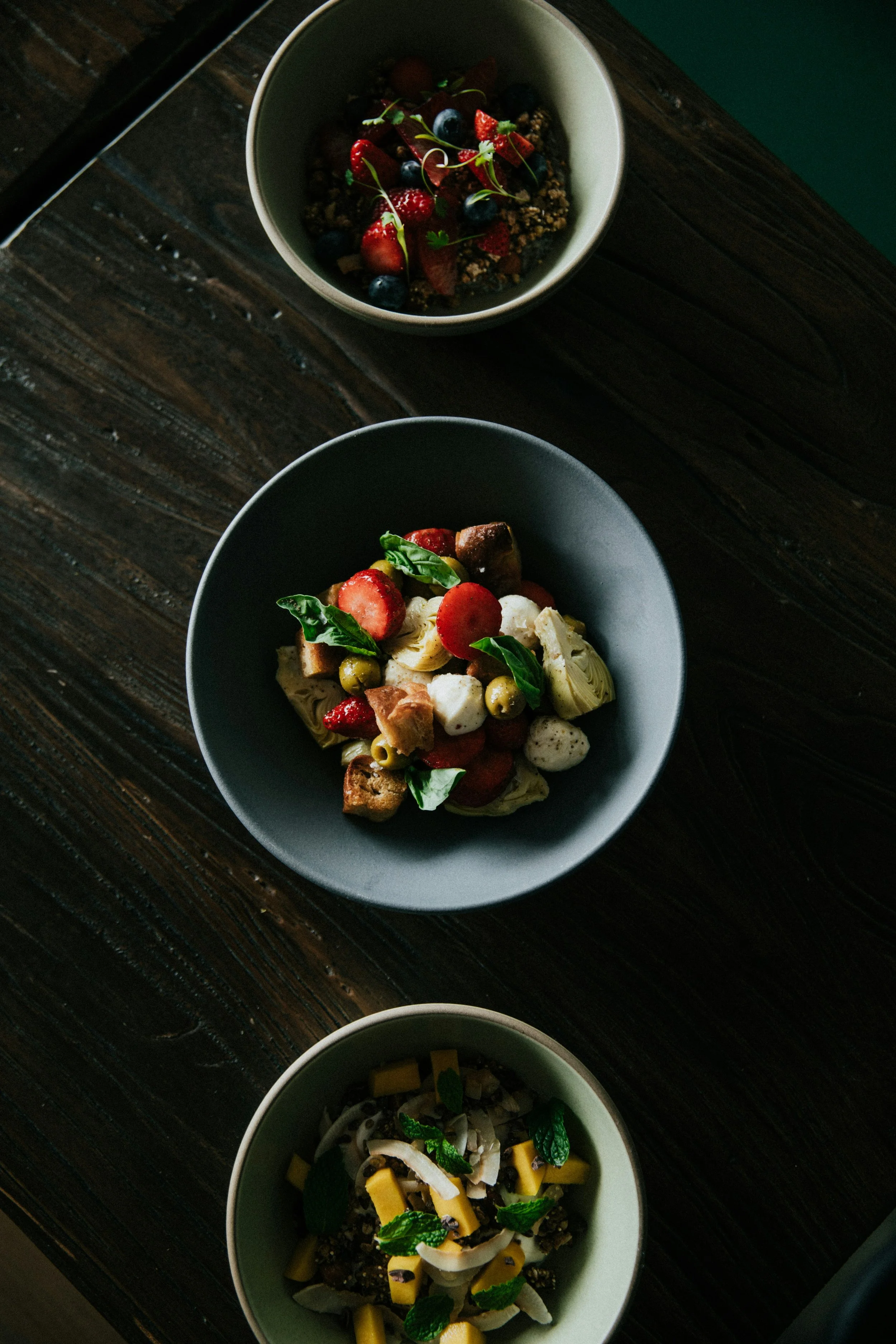 Trois bols de nourriture à base de fruits, légumes et autres ingrédients variés, placés sur une table en bois foncé.