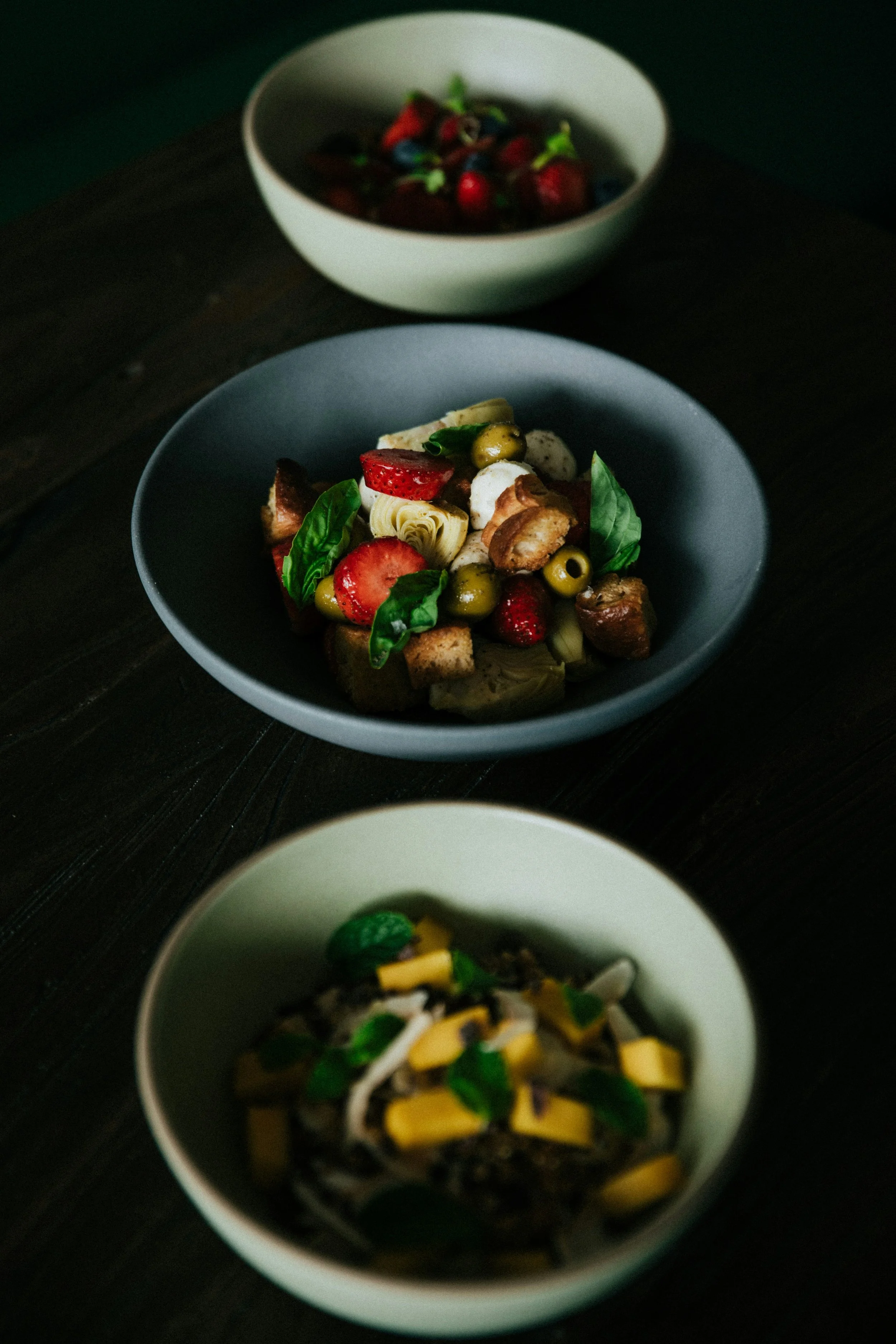 Trois bols de salades diverses avec des légumes et des fruits, placés sur une table en bois foncé