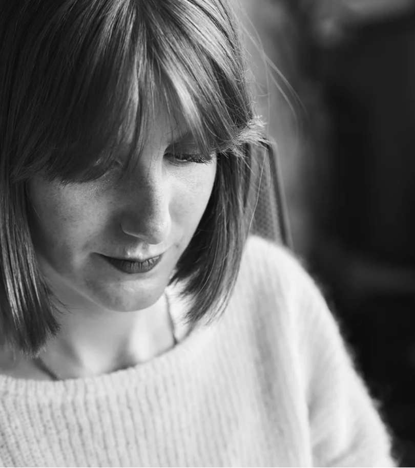 Portrait d'une femme aux cheveux rousse avec une frange, portant un pull blanc, regardant vers le bas, en noir et blanc.