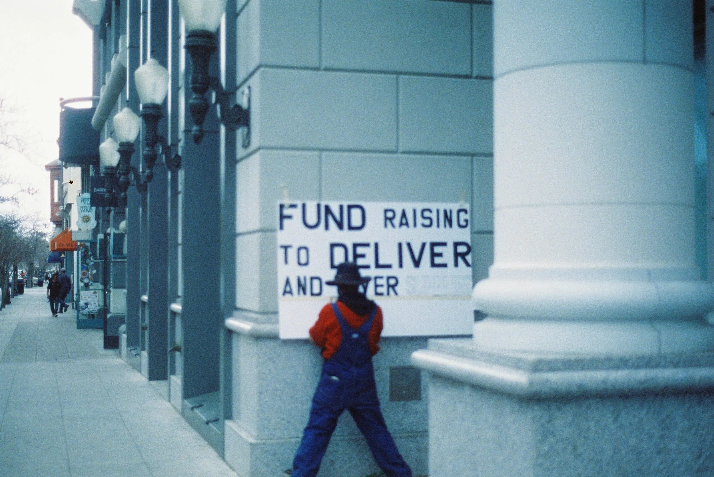 A city sidewalk scene with people walking and a child in a red jacket and blue overalls standing next to a sign that reads 'Fund Raising to Deliver and'.