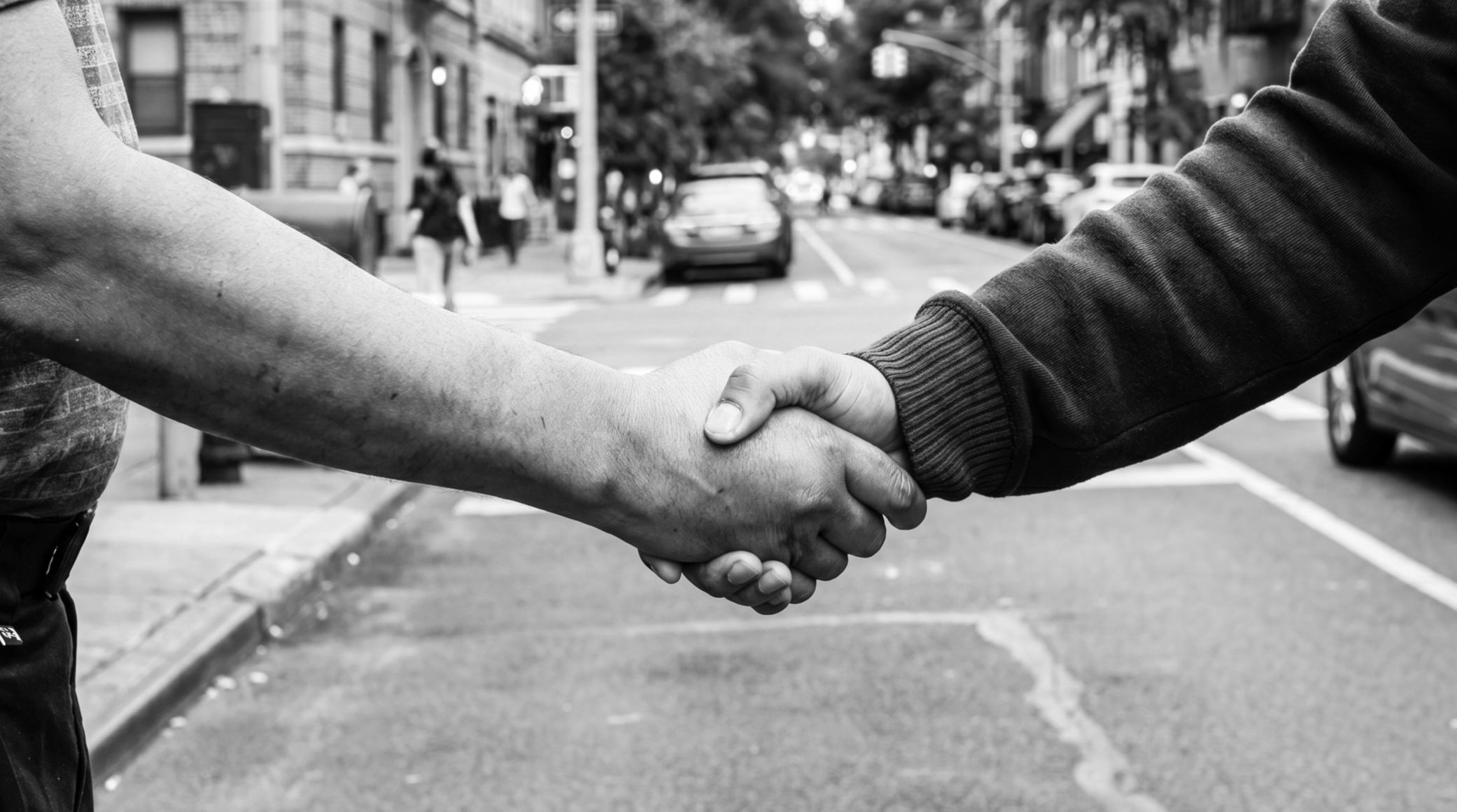 Two people shaking hands on a city sidewalk with cars and pedestrians in the background.