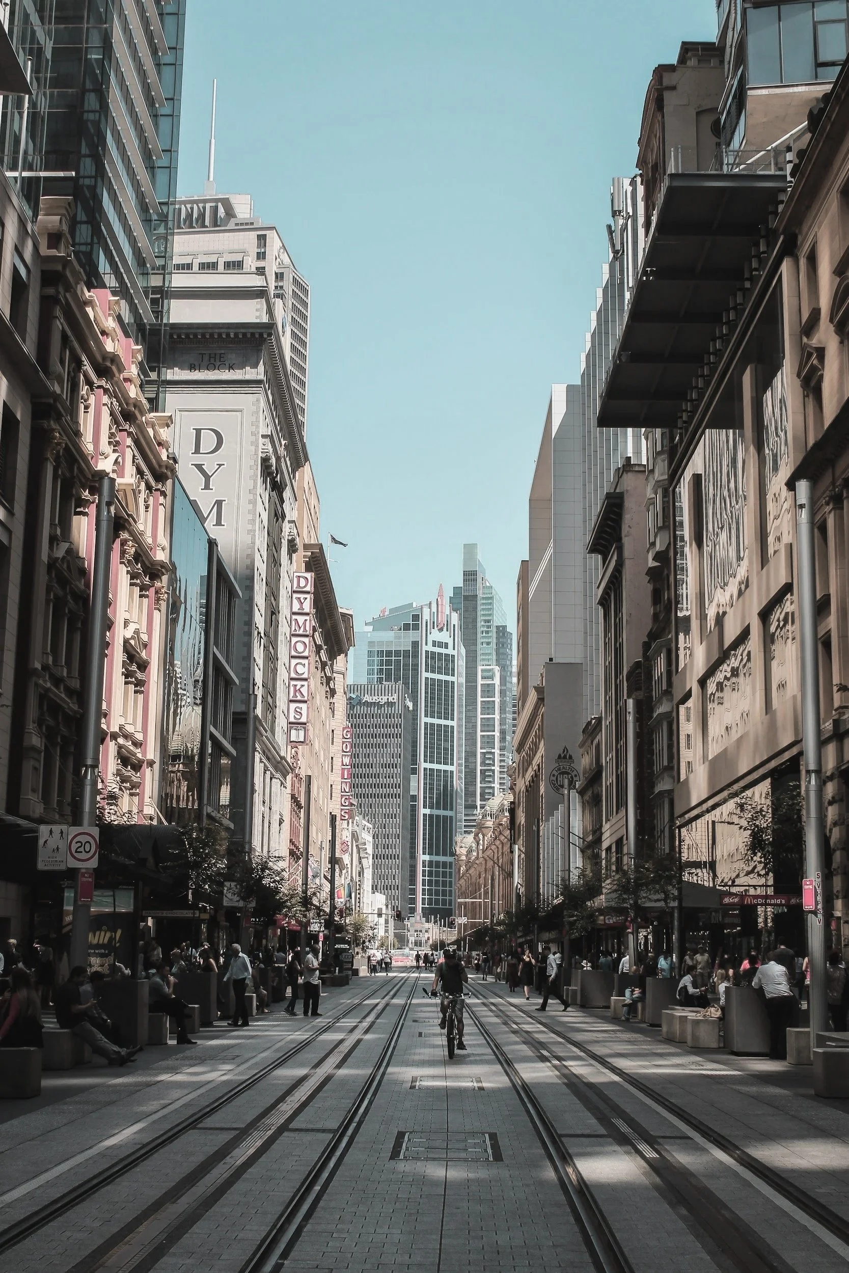 A city street with tall buildings on both sides, people walking, a person riding a bicycle on tram tracks, and signs for shopping and a gym. The sky is clear and blue.