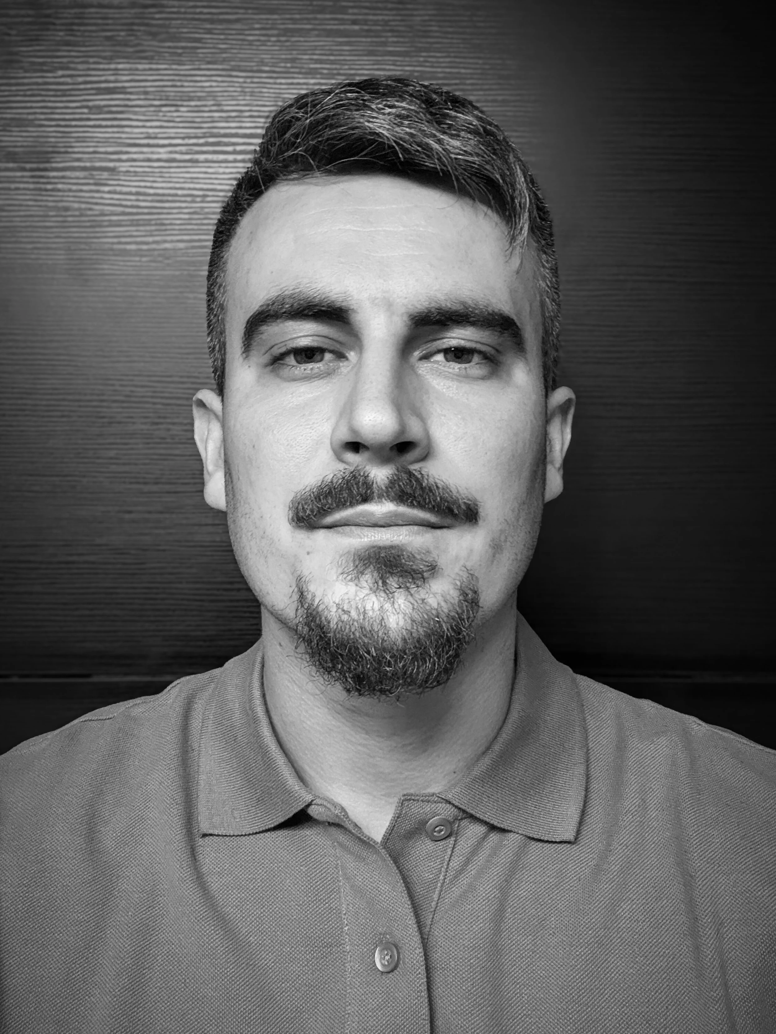 Self-portrait en noir et blanc d'un homme avec une barbe et une moustache, portant une chemise à col, devant un mur en bois.