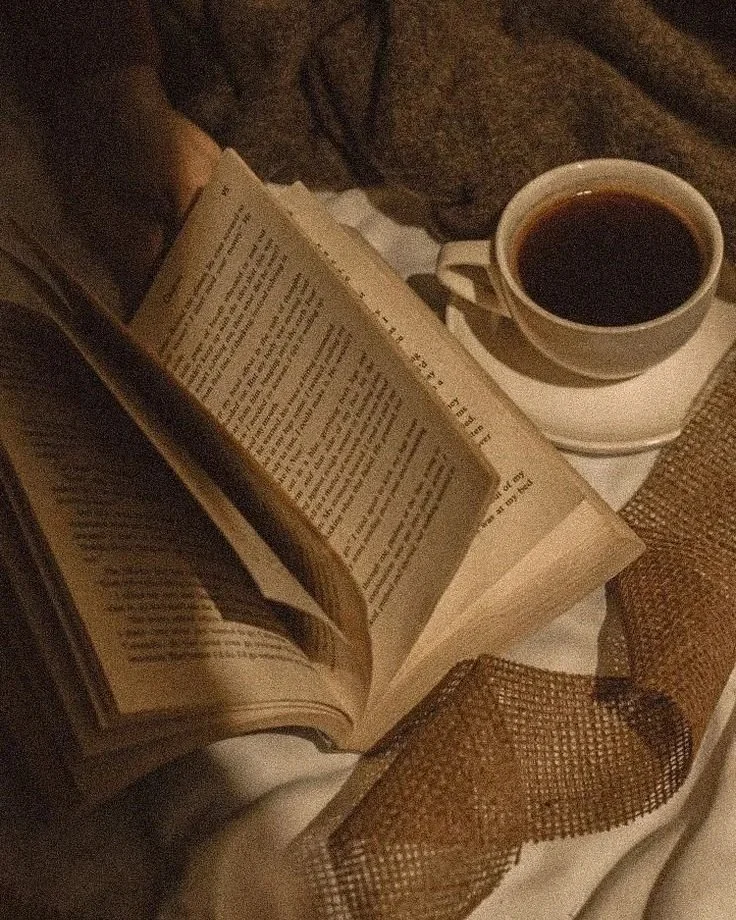 Open book, cup of coffee on saucer, brown mesh ribbon, blanket or sweater in the background on a white surface.