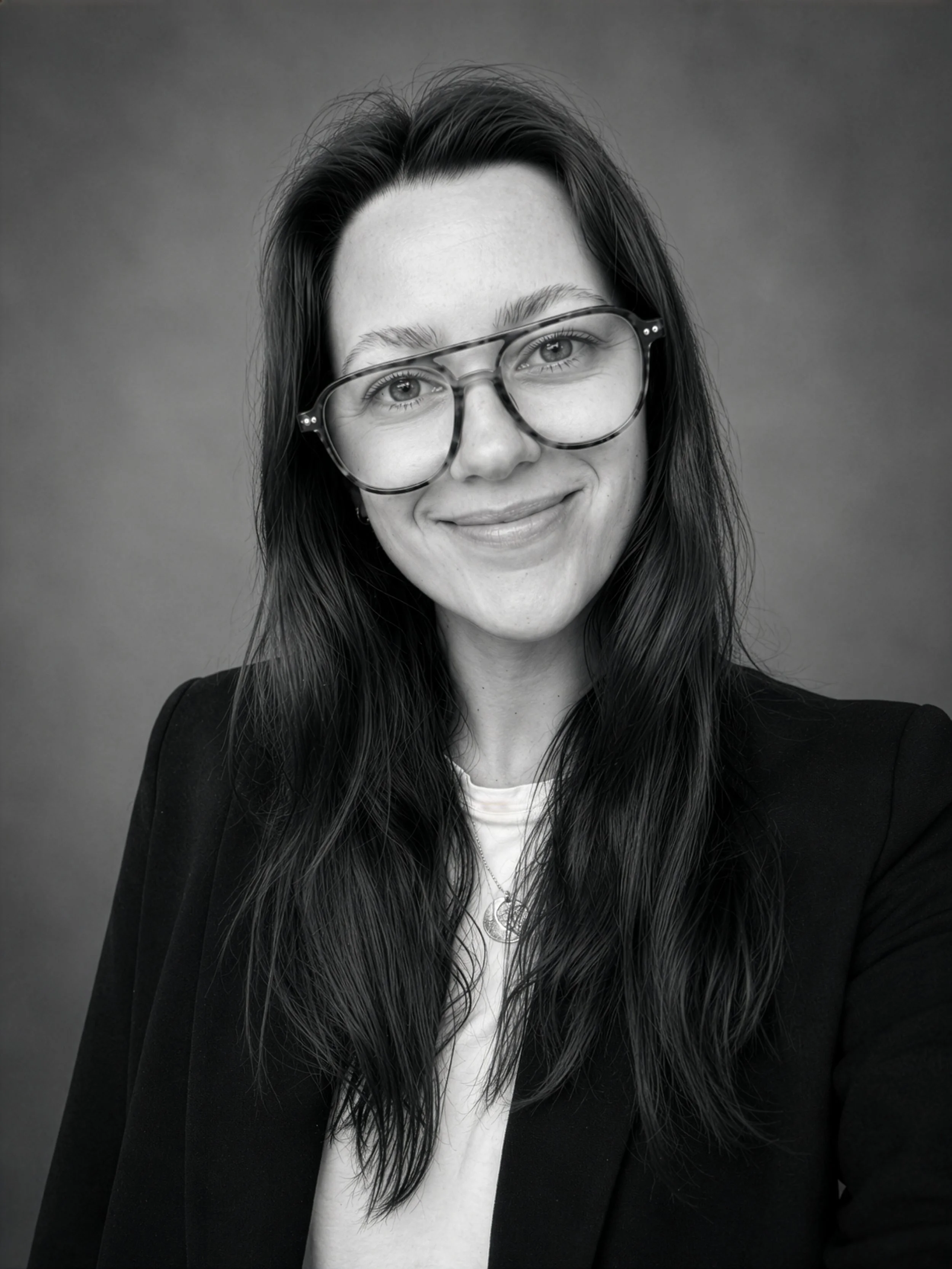 A young woman with long dark hair, wearing large tortoiseshell glasses, a white top, and a black blazer, smiling at the camera against a plain background.