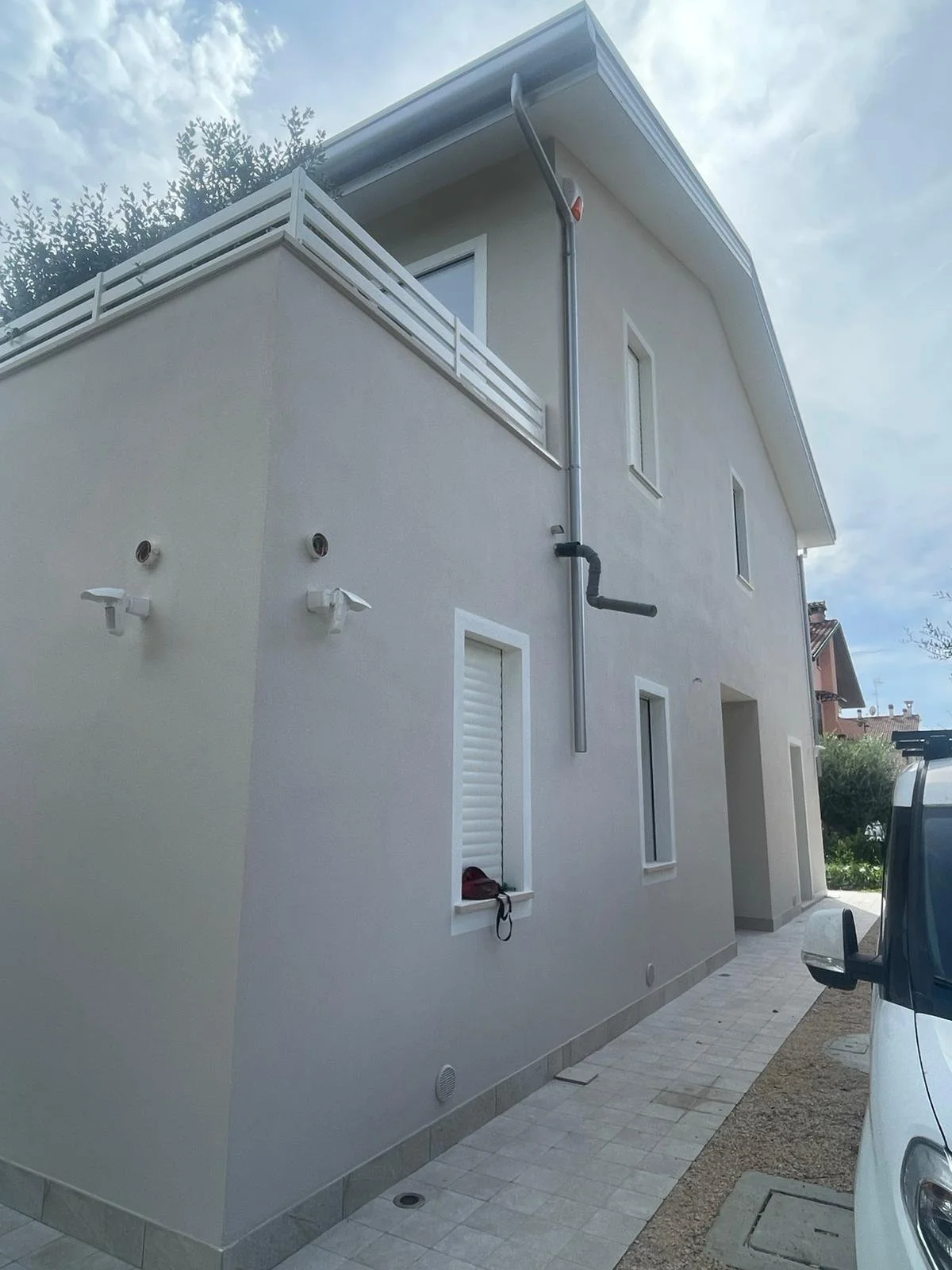 Side of a modern, light-colored house with window shutters, a balcony, and external pipes, set against a partly cloudy sky.