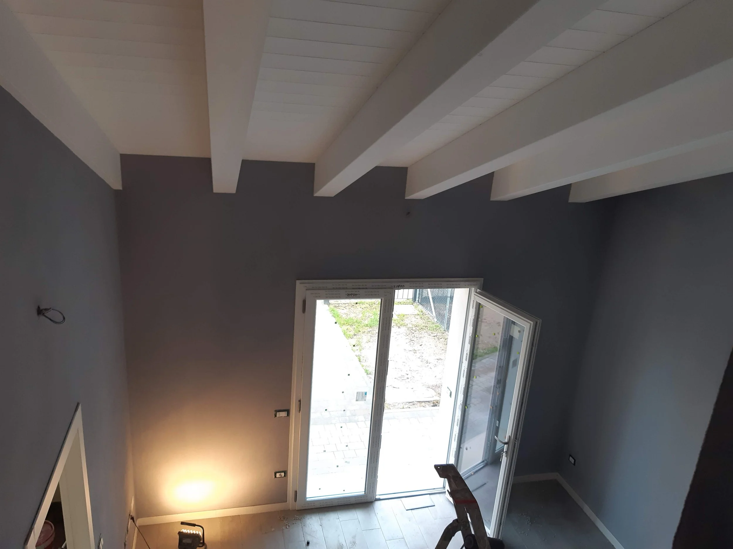 A room under construction with a large sliding glass door leading outside, unfinished flooring, and unpainted walls. Natural light enters through the glass door, illuminating part of the room.