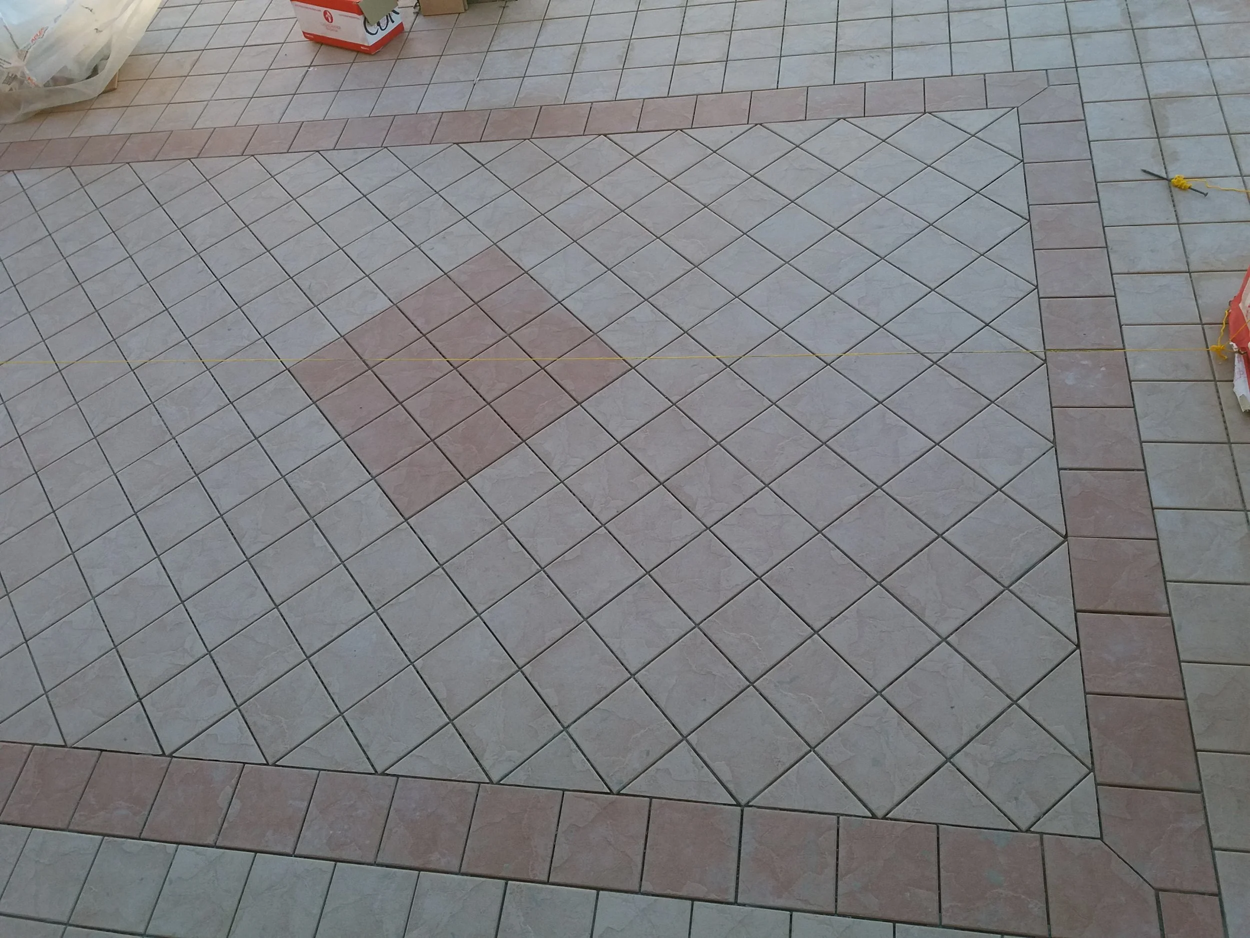 Close-up view of a tiled floor with square tiles in beige, pink, and light green colors arranged in a pattern. There are some construction or renovation items, including a plastic bag, a red box, and yellow string, visible on the floor.