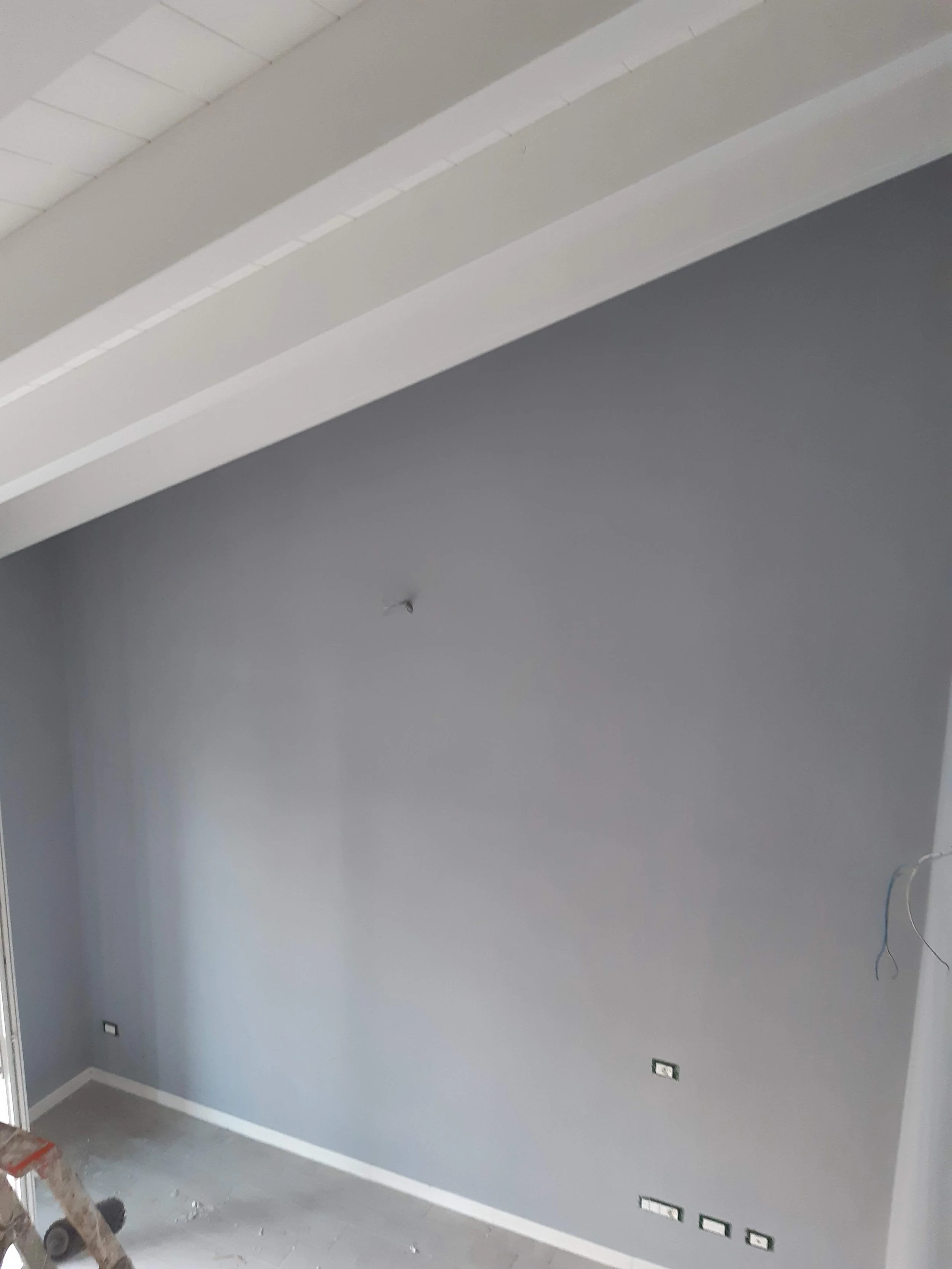 Interior of a room with gray walls, white ceiling with panel tiles, electrical outlets on the wall, and some construction tools and materials in the corner.