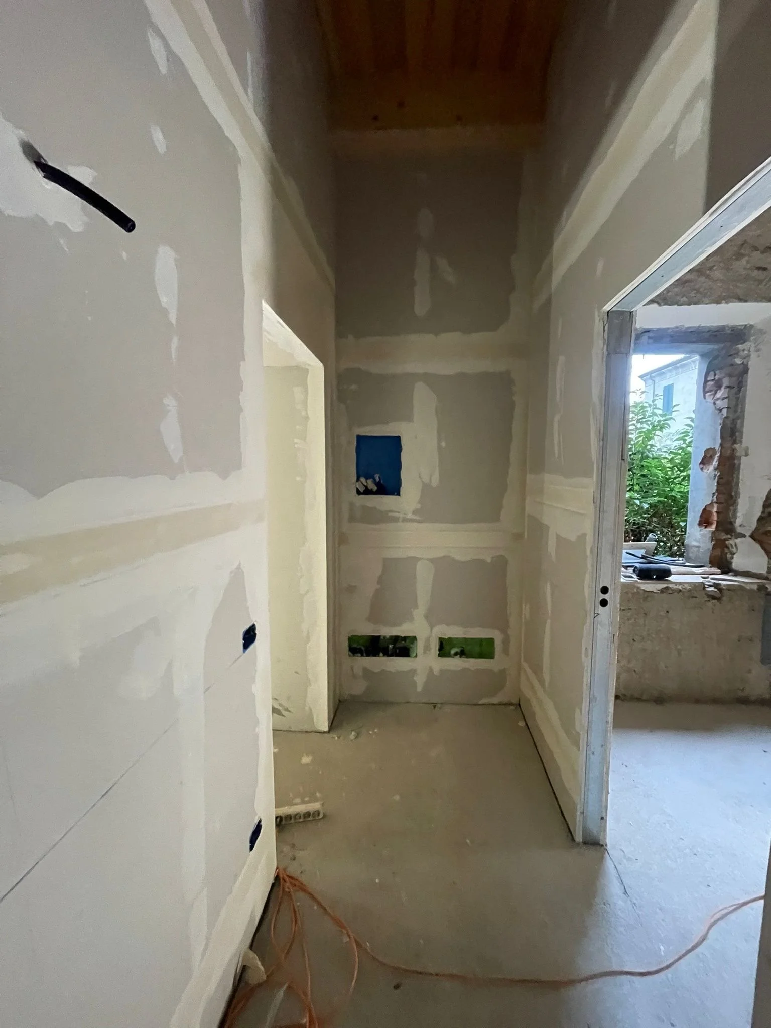 Interior of a room under construction with partially finished drywall walls, an opening to another room, and construction tools and wires on the floor.