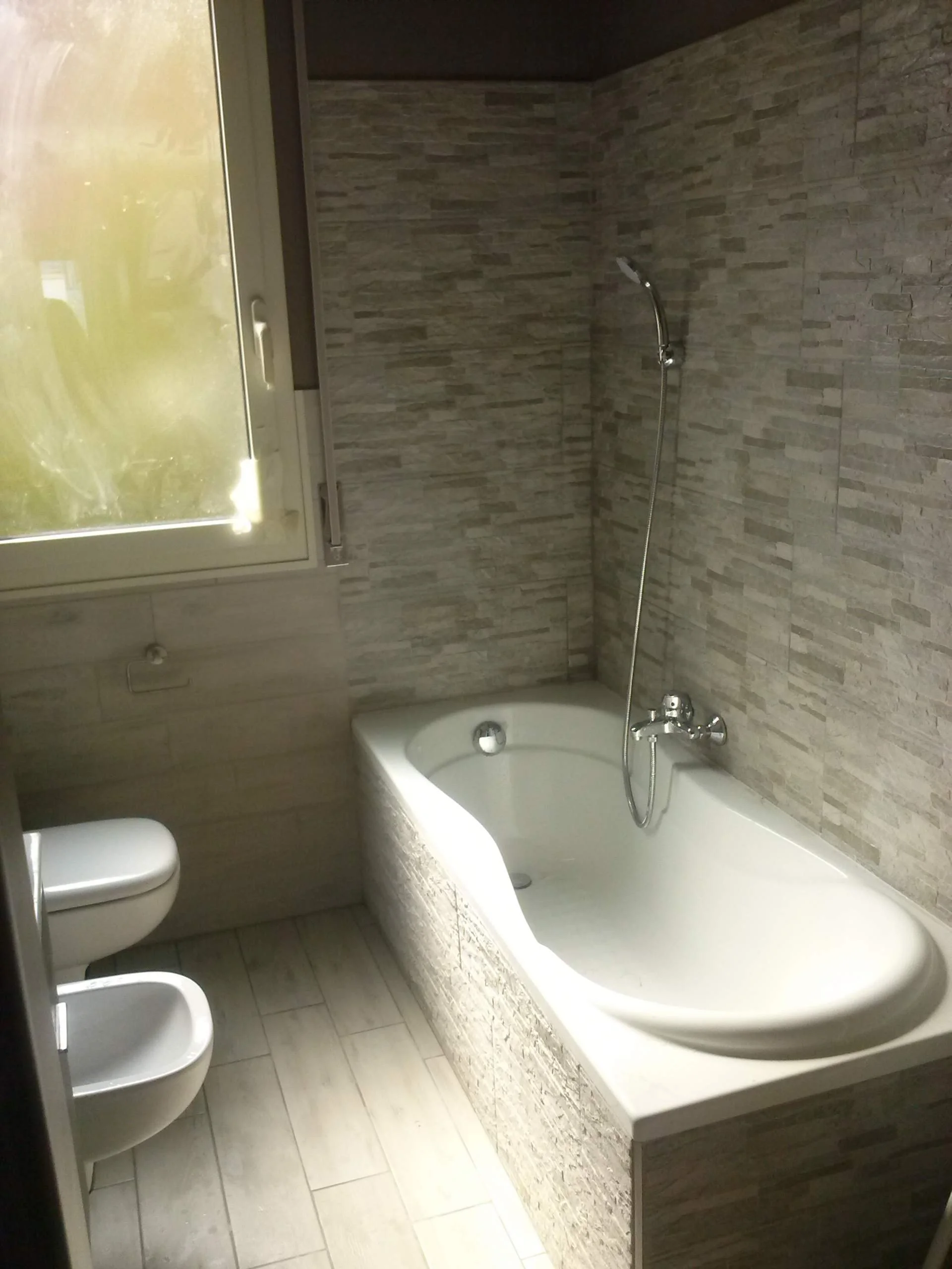 Bathroom with bathtub, toilet, bidet, and window. Tiled walls and floor.