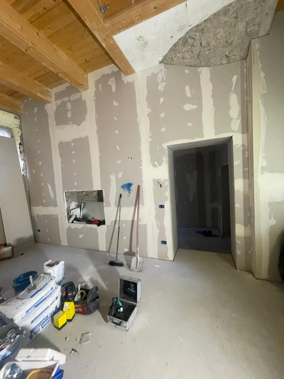 Interior of a house under construction with unfinished drywall, wooden ceiling beams, and construction tools and materials scattered on the floor.