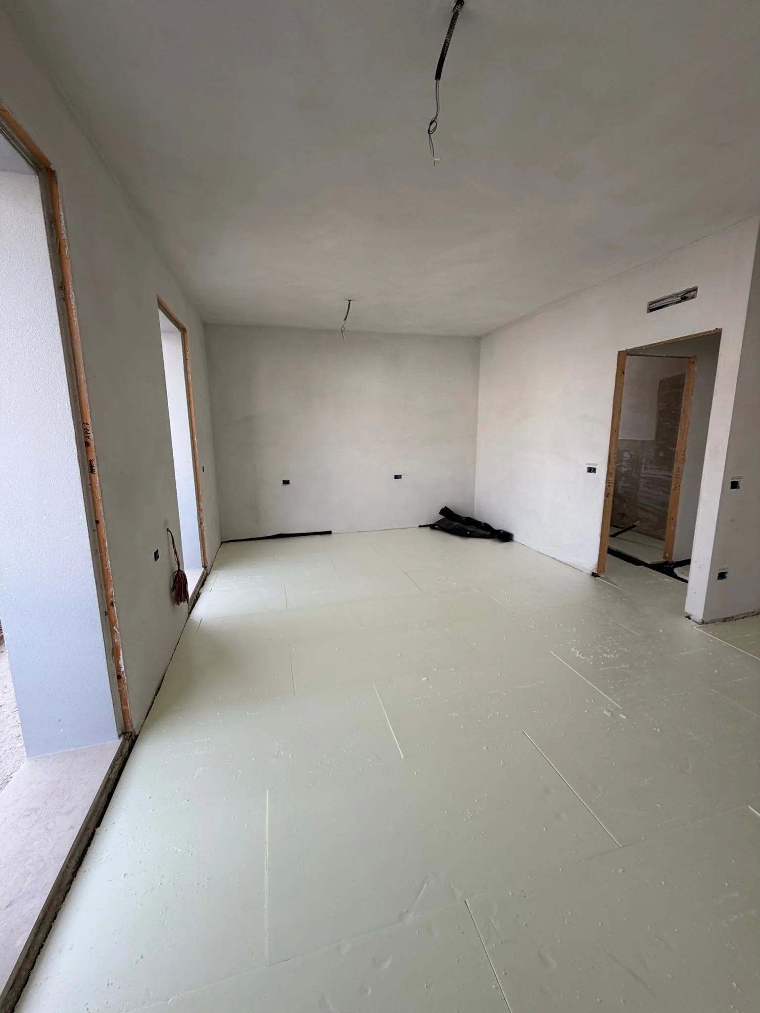 Interior of a room under construction with unfinished walls and ceiling, electrical wiring hanging from the ceiling, and construction materials on the floor.