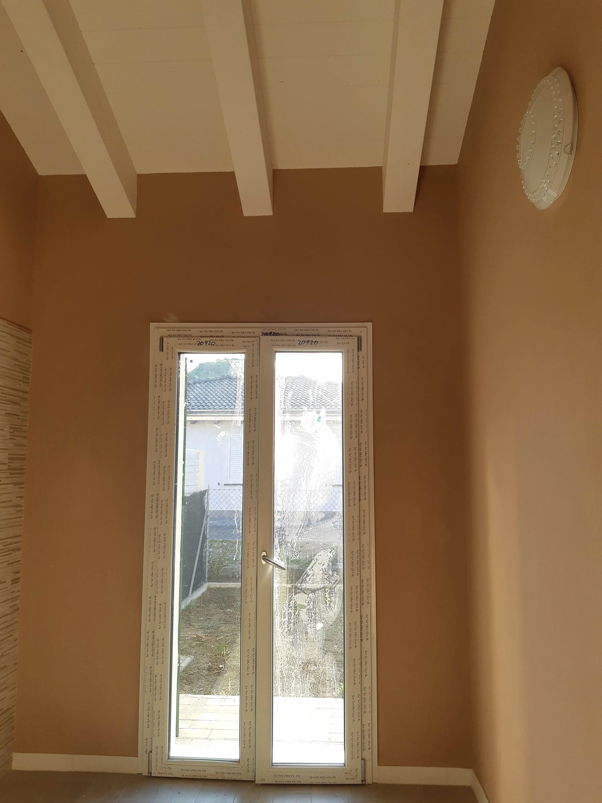 Interior room with large window and sliding glass door leading outside, with a view of a house with tiled roof and yard.