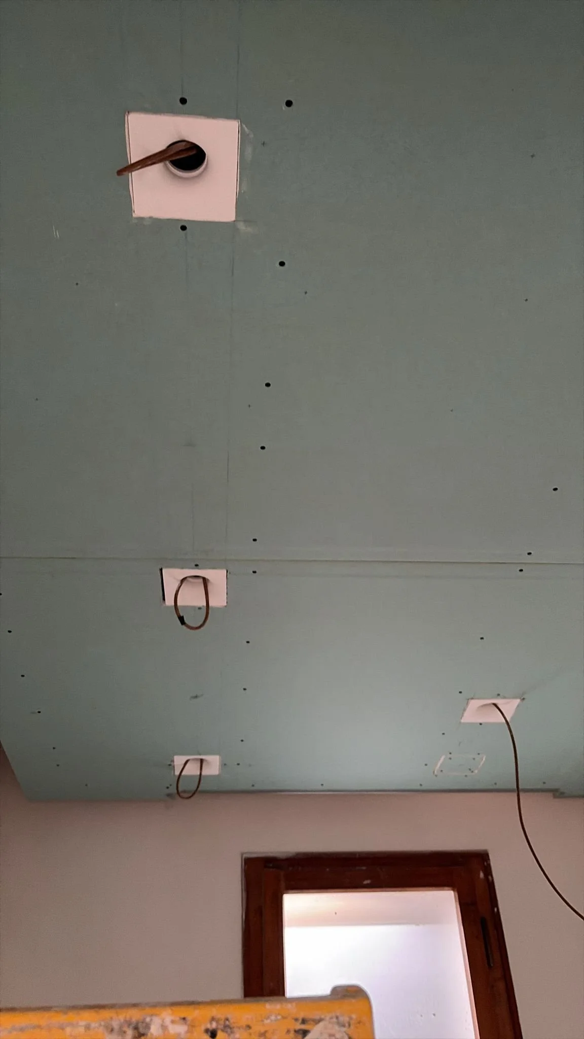 Ceiling with multiple electrical outlets, some with hanging wires, and a window with a wooden frame.