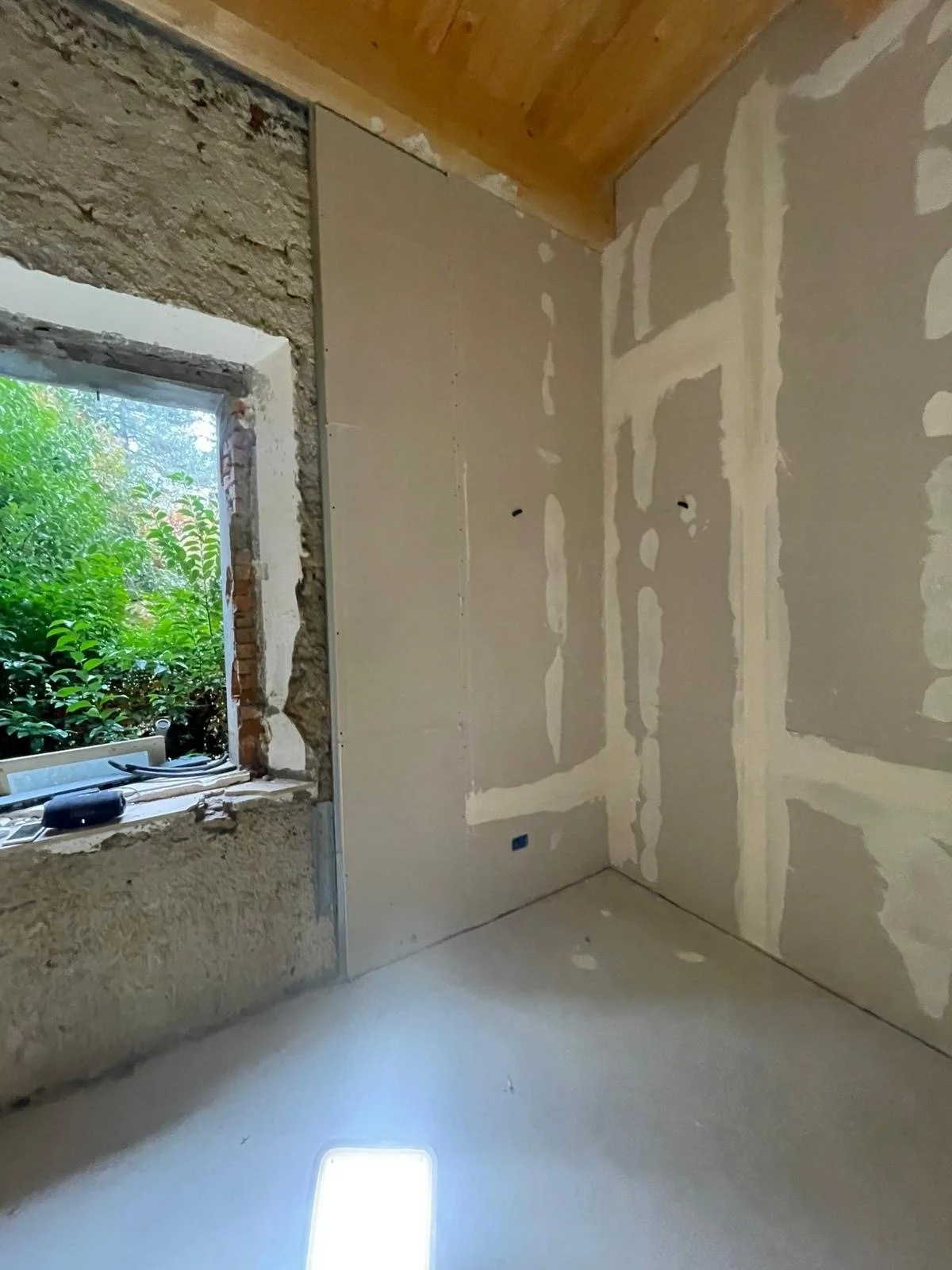An interior room under construction with drywall partially installed. The wall on the right has visible joint compound and taped seams. A window on the left shows green foliage outside, with some construction materials on the windowsill.