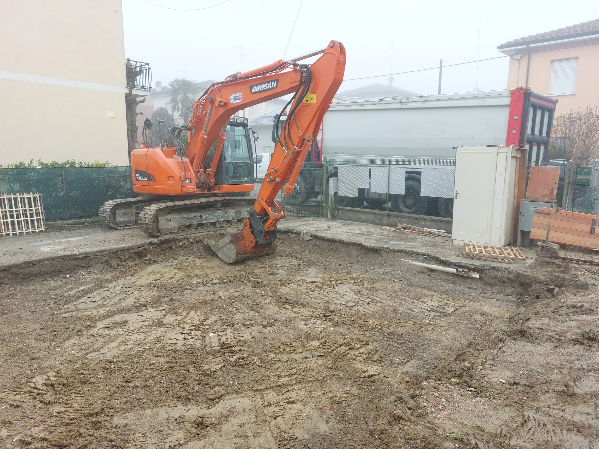 A construction site with an orange Doosan excavator, dirt ground, and residential buildings in the background, partially obscured by fog.