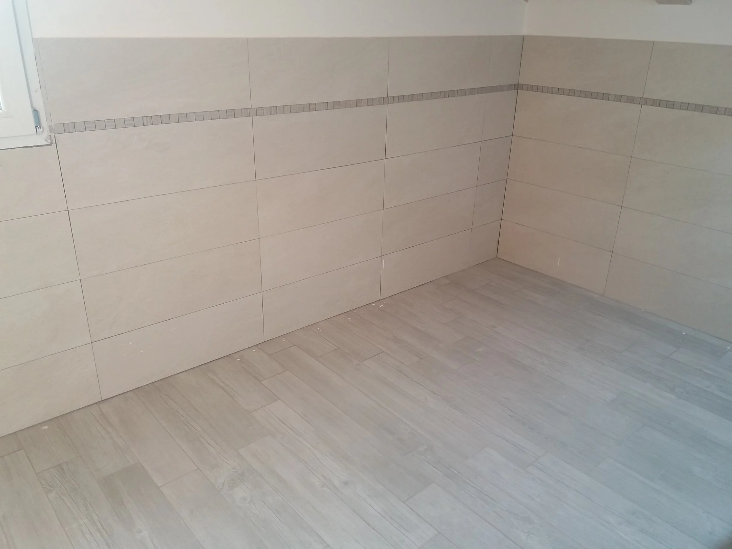 Interior of a room with tiled walls and wooden flooring, light-colored tiles with a decorative border, and a small window on the left.