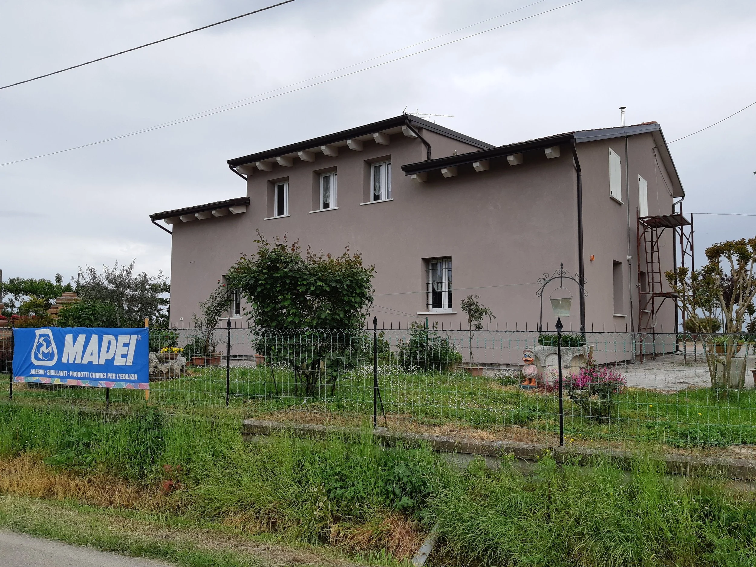A two-story house with a beige exterior, multiple windows, and a shed roof, surrounded by a garden with flowers, trees, and a black metal fence. There is a blue banner with the logo 'MAPEI' in front of the fence.