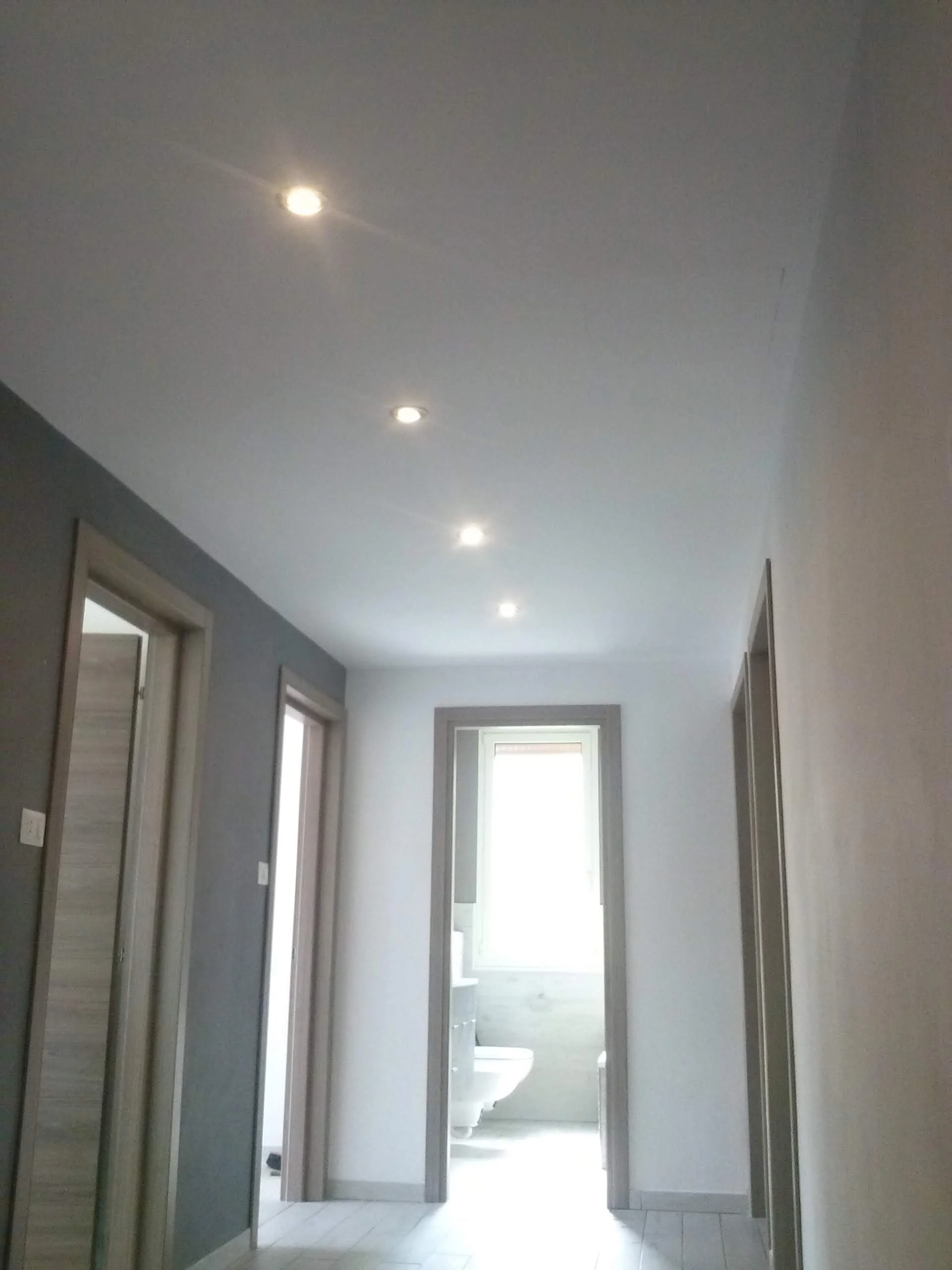 A hallway with four doorways, the central door leading to a bathroom with a white toilet visible, and natural light streaming in from a window at the end of the hallway.