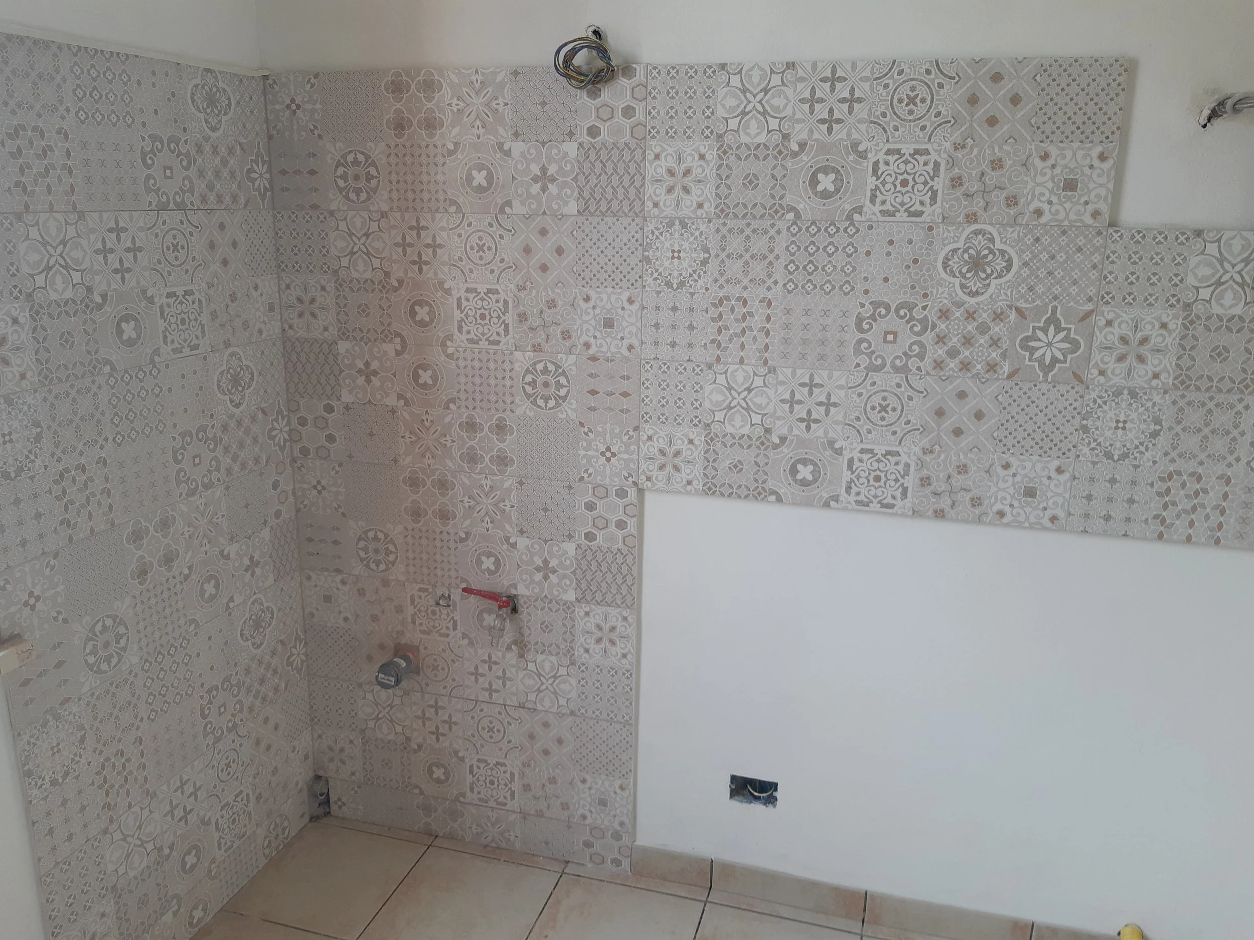 A room under construction with partially tiled wall featuring various patterned tiles and an electrical outlet without cover.