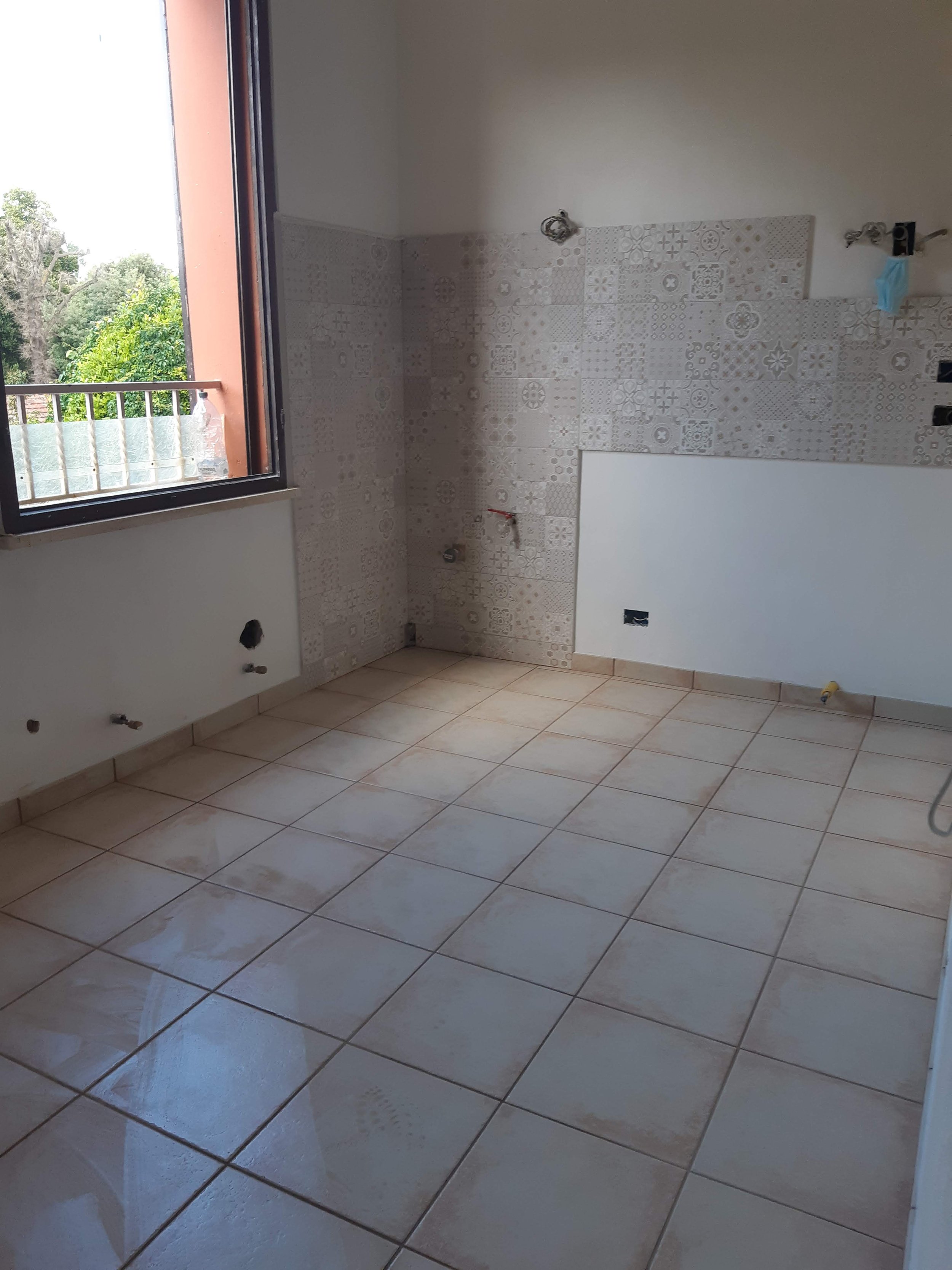 Empty room with tiled floor and a window with a view of trees outside, partially tiled wall, electrical outlets, and plumbing fixtures.