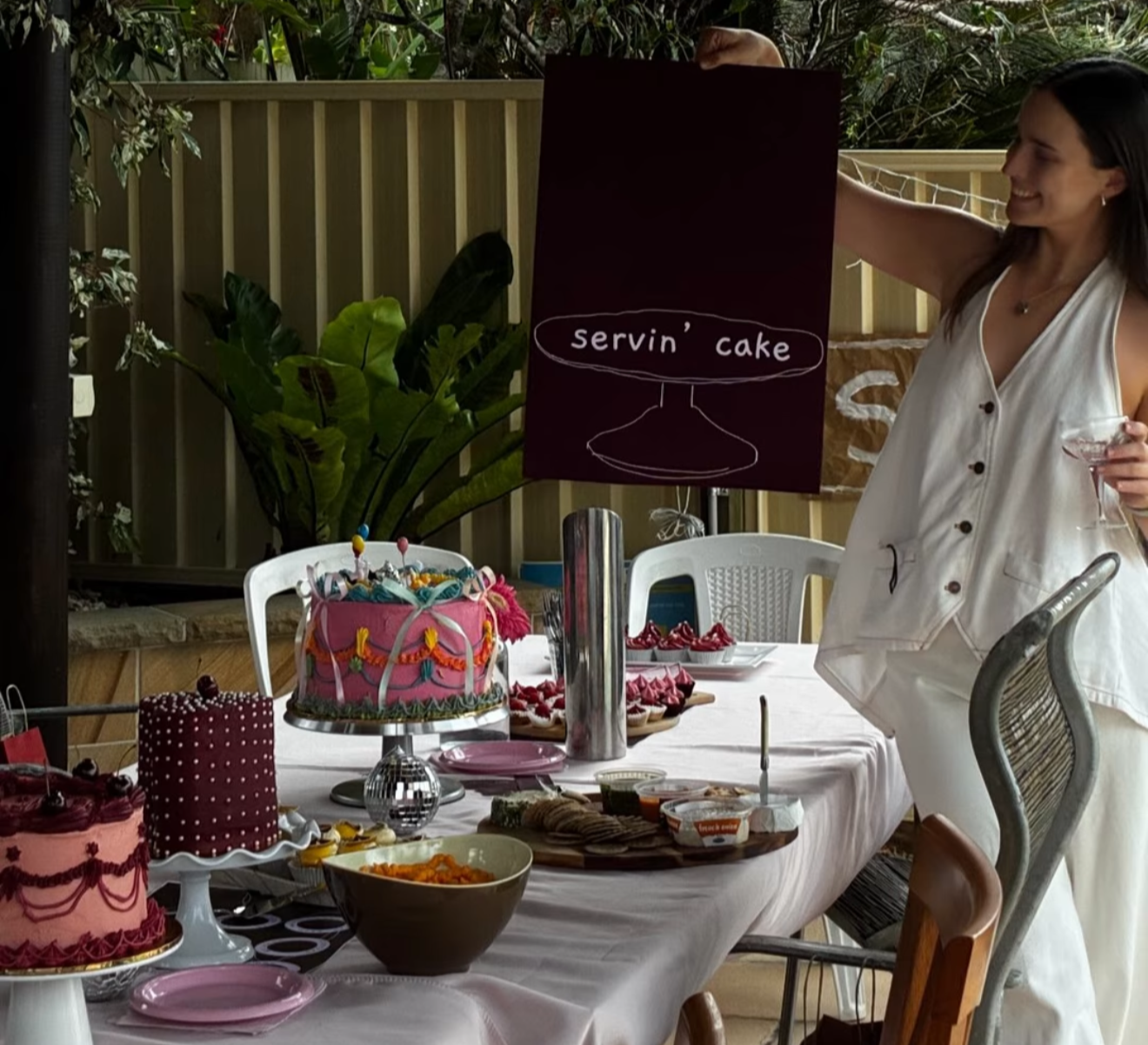 A woman standing next to a table filled with different cakes and desserts during a celebration, with a sign behind her that says 'Servin' cake'.