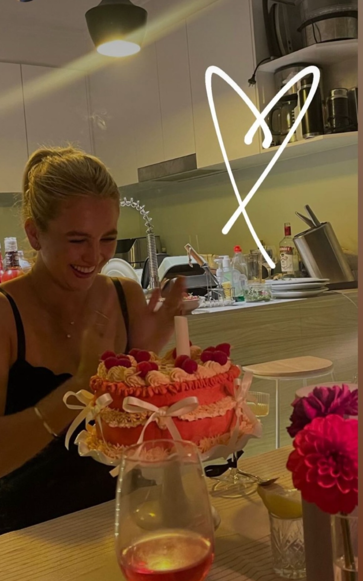 A woman with blonde hair, smiling and waving, celebrates a birthday with a decorated cake topped with raspberries, and a wine glass in front of her. The background shows a kitchen with various appliances and dishes.