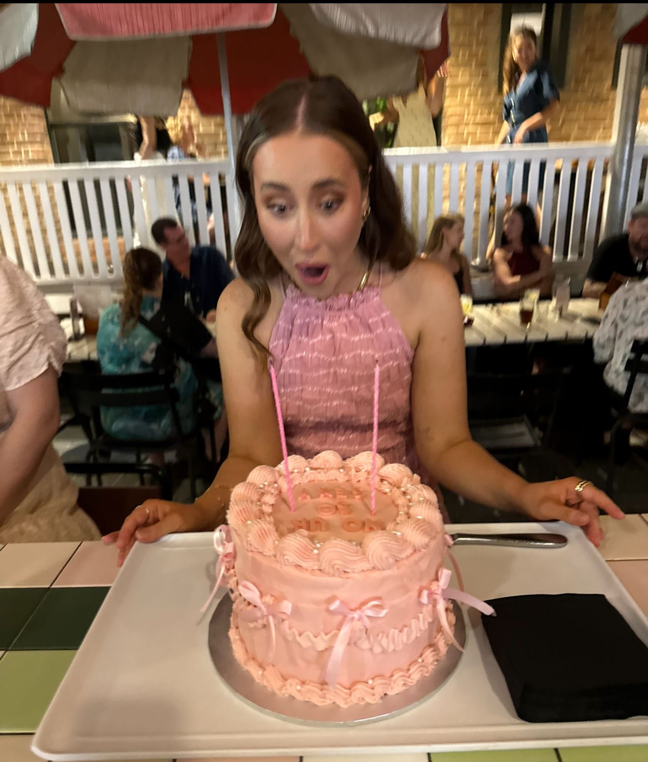 A woman with brown hair in a pink dress is surprised as she looks at a pink birthday cake in front of her at a celebration.