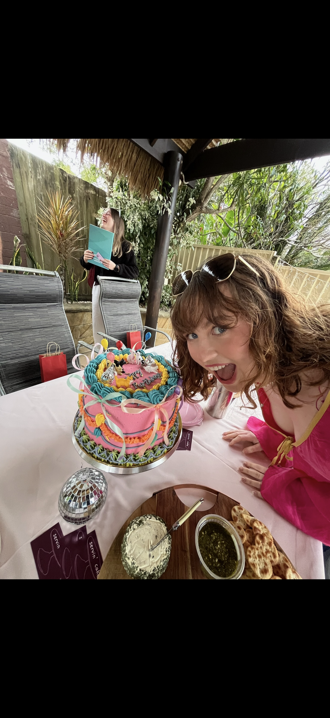A woman in a pink dress and sunglasses on her head leans forward and smiles with her mouth open at a colorful birthday cake on a table. In the background, a woman stands holding a book and laughing, while a festive outdoor setting features chairs, a wooden table with food, and lush green plants.