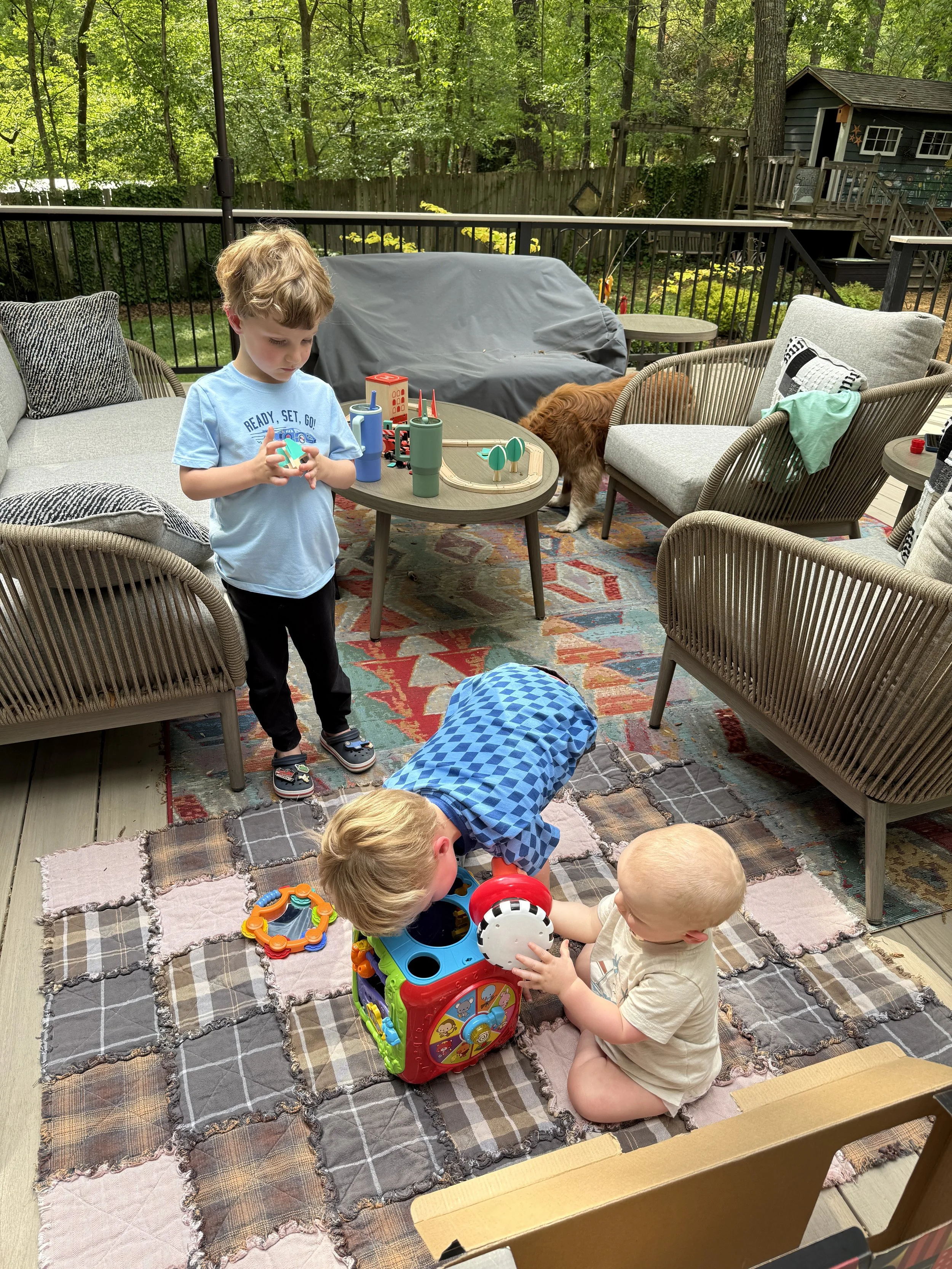 Children playing with toys on a porch with outdoor furniture, surrounded by trees and a backyard.
