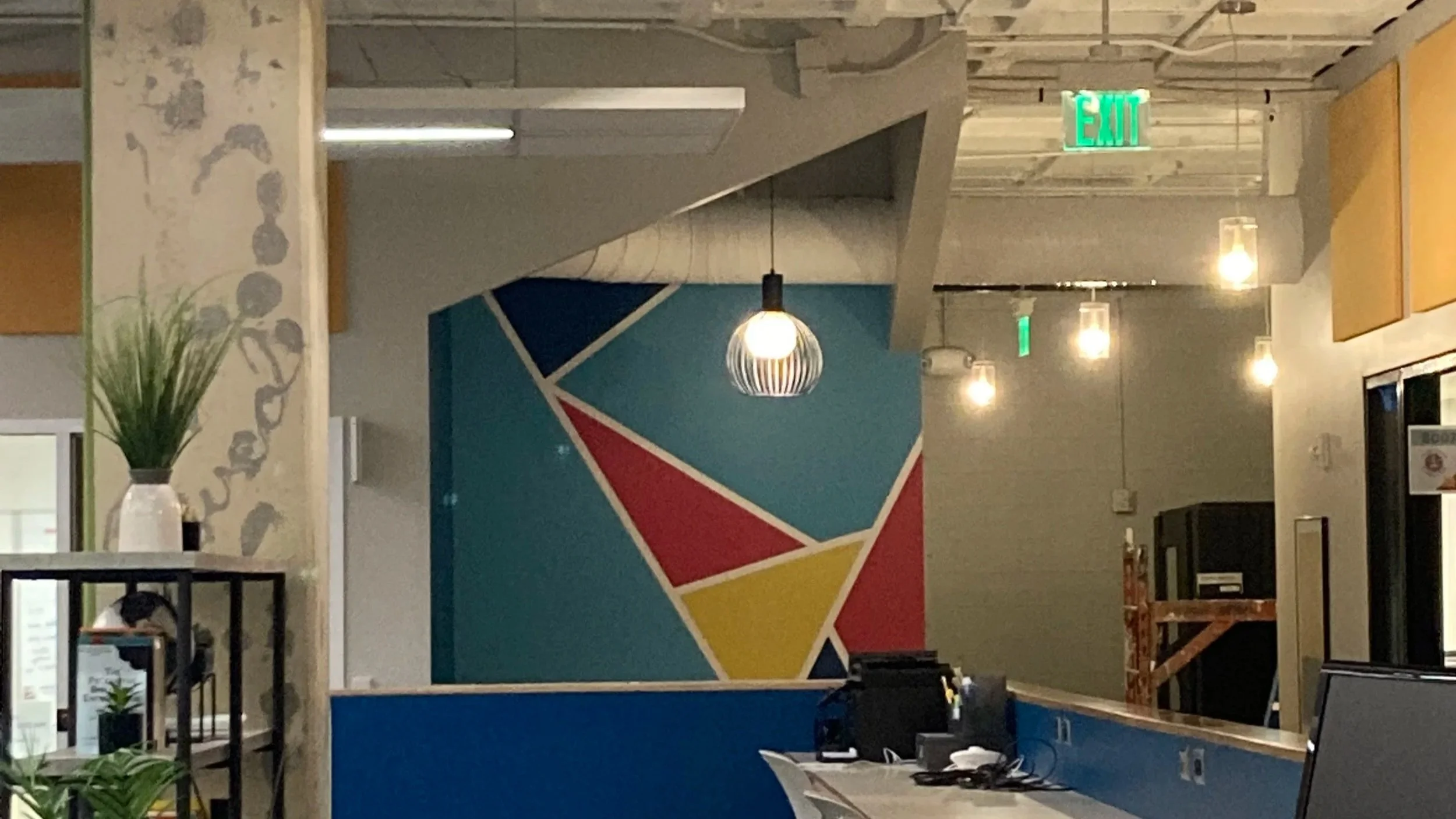 Interior of American Underground, a modern office co-working space featuring a colorful geometric mural, hanging pendant lights, plants, and various office furniture.